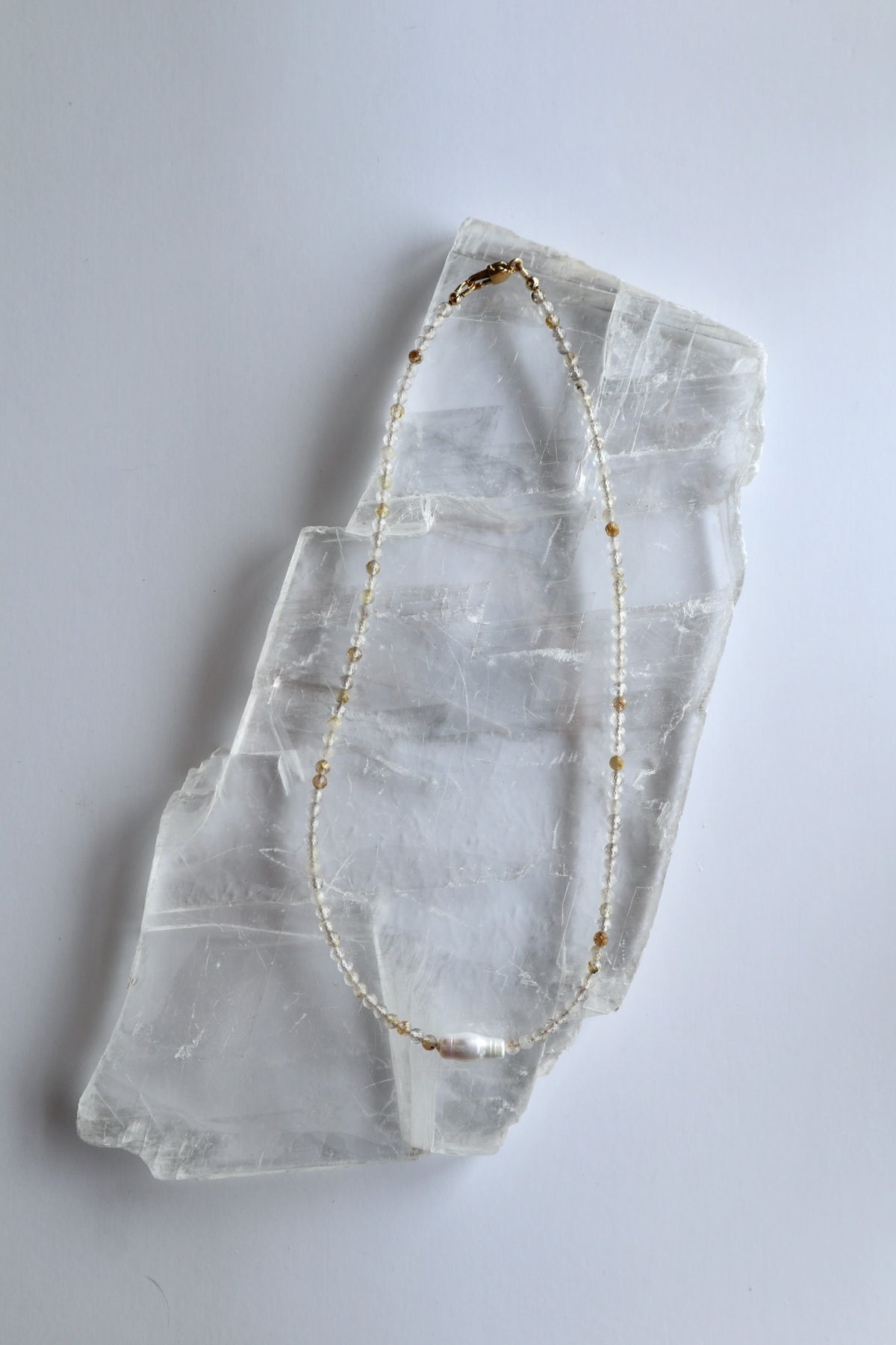Larch necklace by Auréad Designs laid on a white selenite slab, full view of the delicate golden rutilated quartz strand with single baroque freshwater pearl focal bead, Glacier Collection