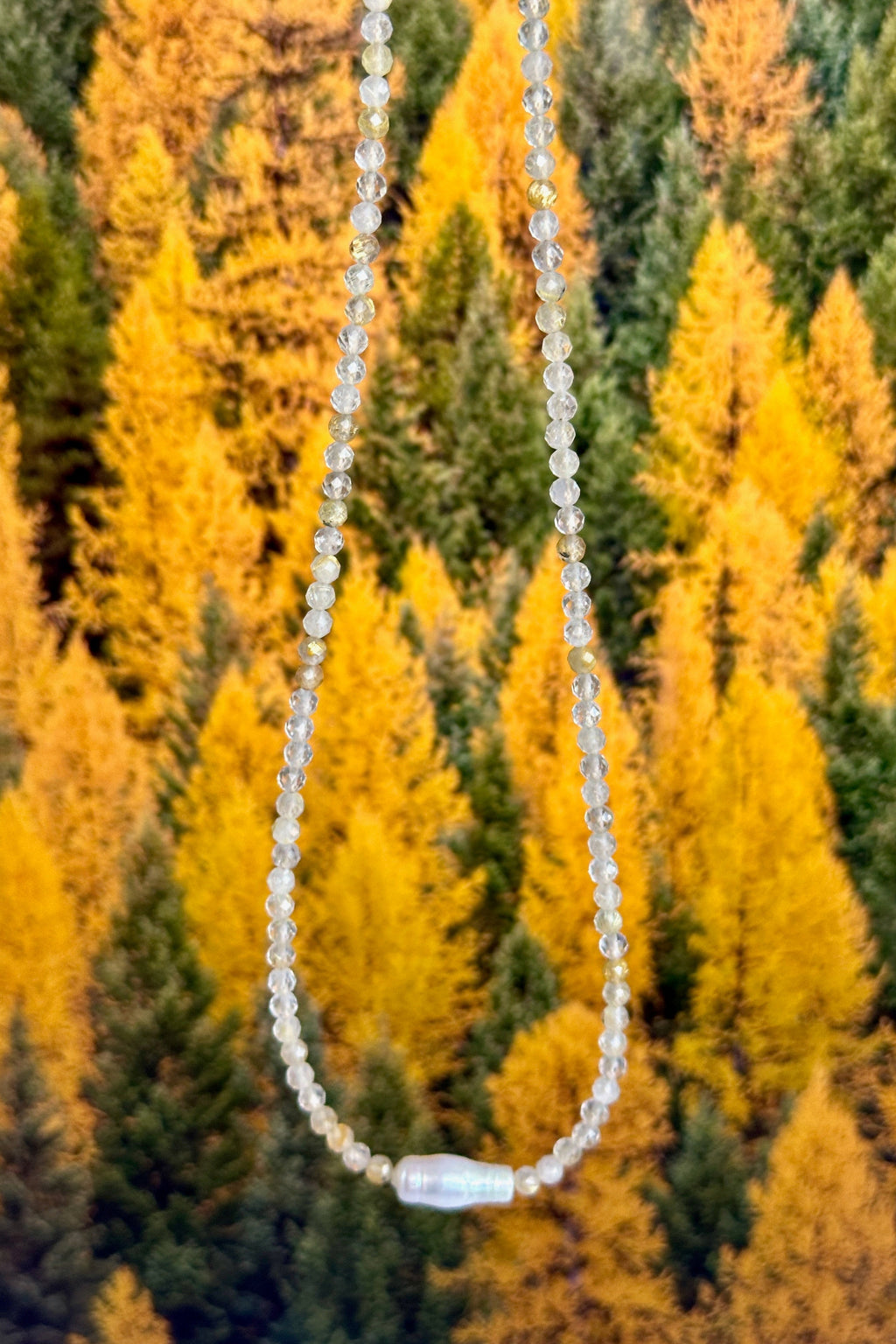Larch necklace by Auréad Designs held against golden larch trees along the Icefields Parkway, showing the full golden rutilated quartz strand with baroque freshwater pearl focal bead, Glacier Collection
