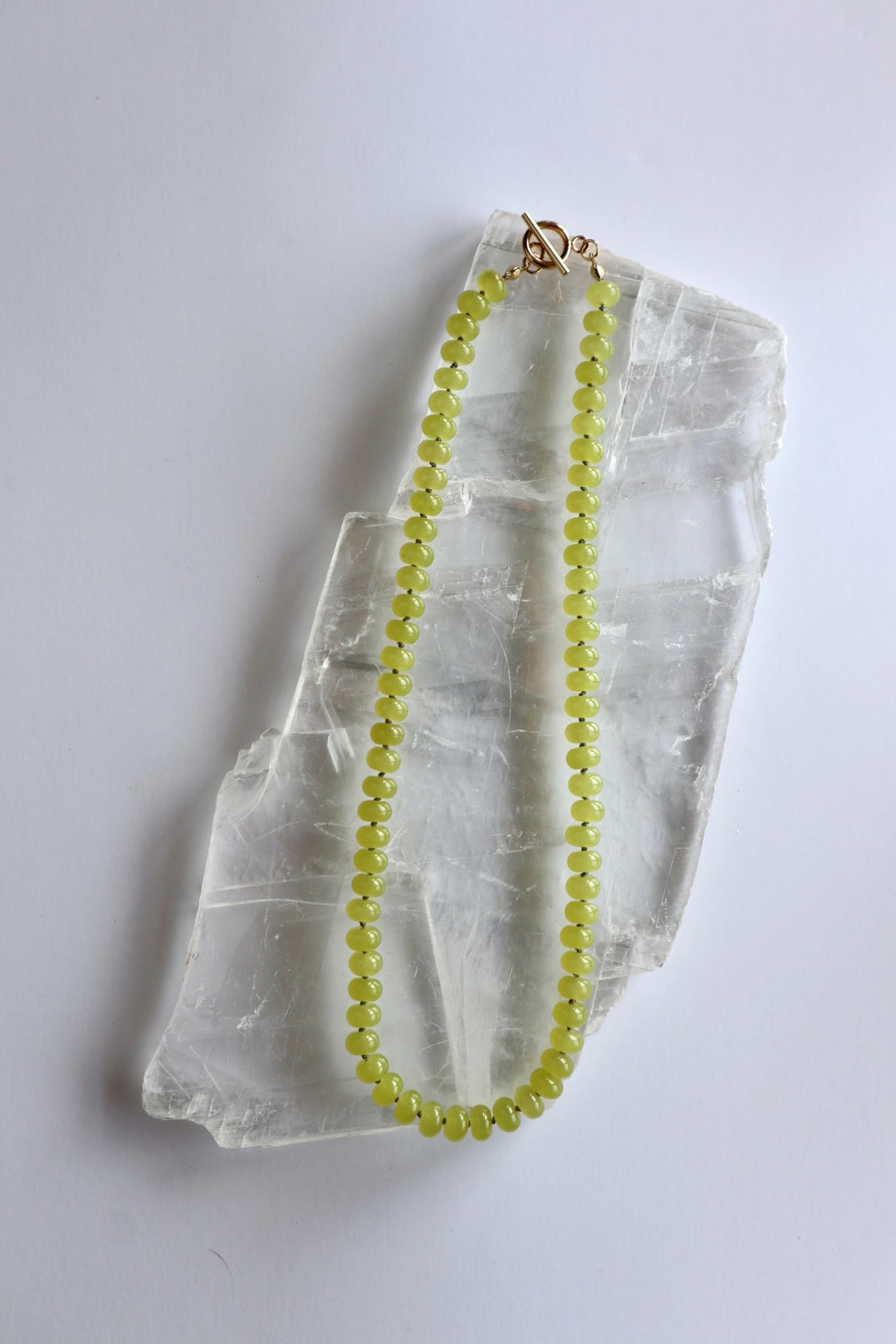 Ledge necklace by Auréad Designs laid on a white selenite slab, full view of the lemon jade rondelle strand with gold toggle clasp, Glacier Collection