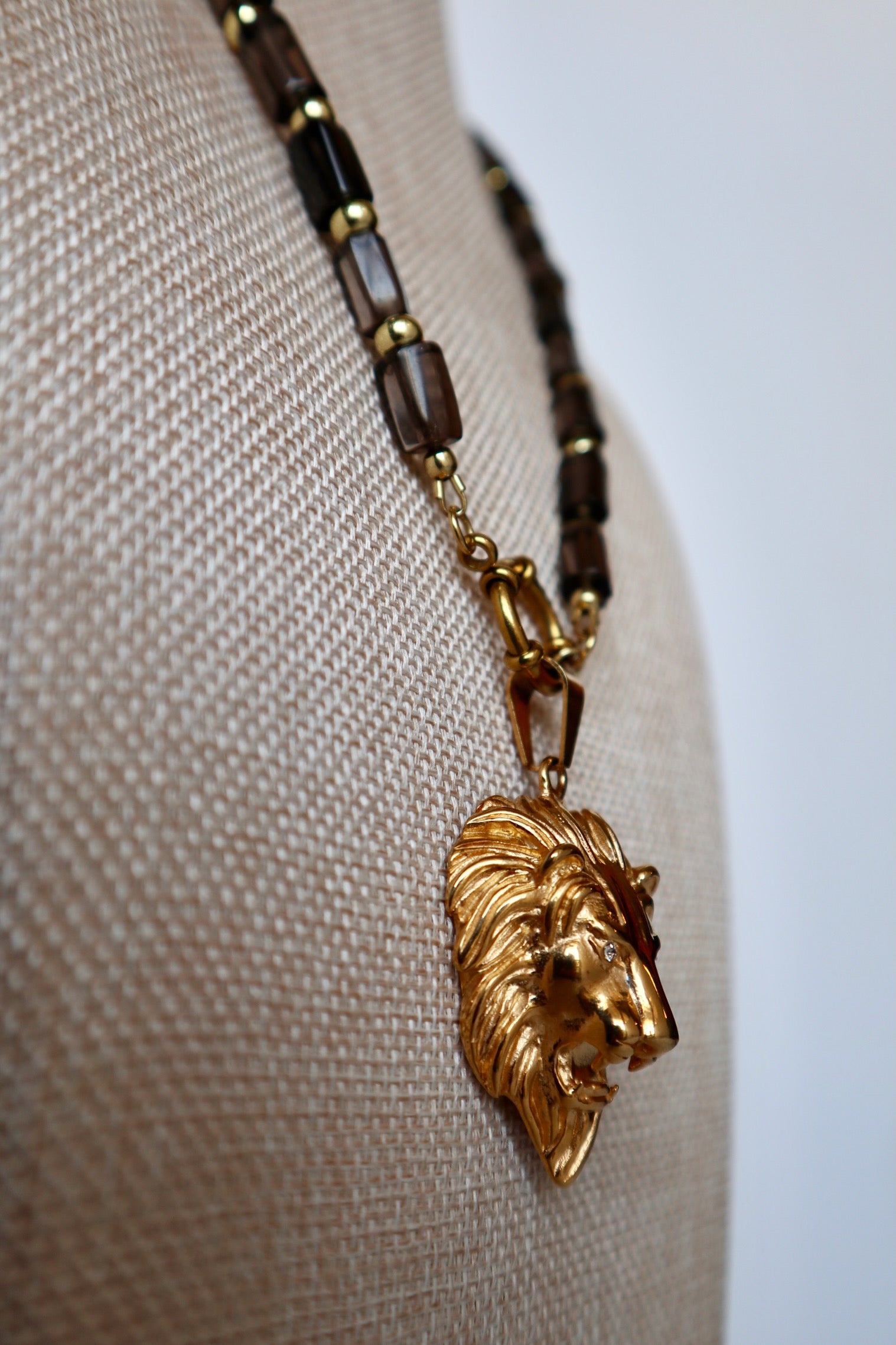 Auréad Designs Leoven necklace draped at an angle on linen bust form, showing smoky quartz cylinder beads, gold spacers, and gold lion head pendant with spring ring clasp
