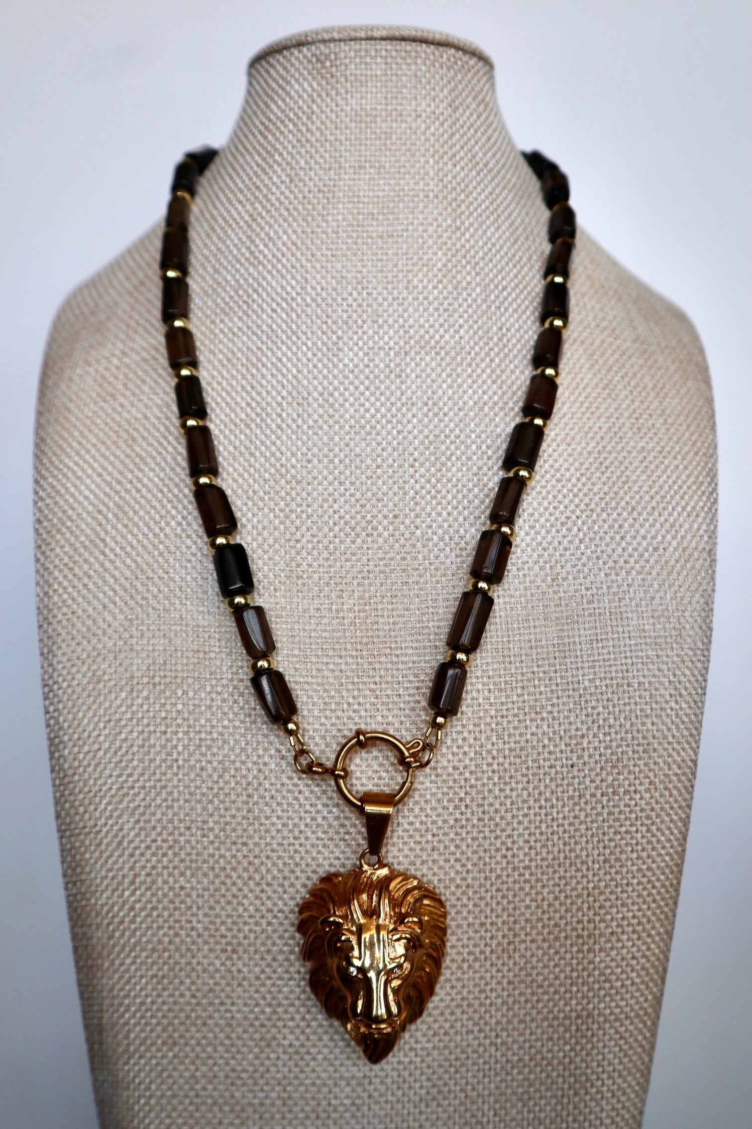 Auréad Designs Leoven necklace displayed front-facing on linen bust form, showing faceted smoky quartz cylinder bead strand with gold spacers and gold lion head pendant centered at front