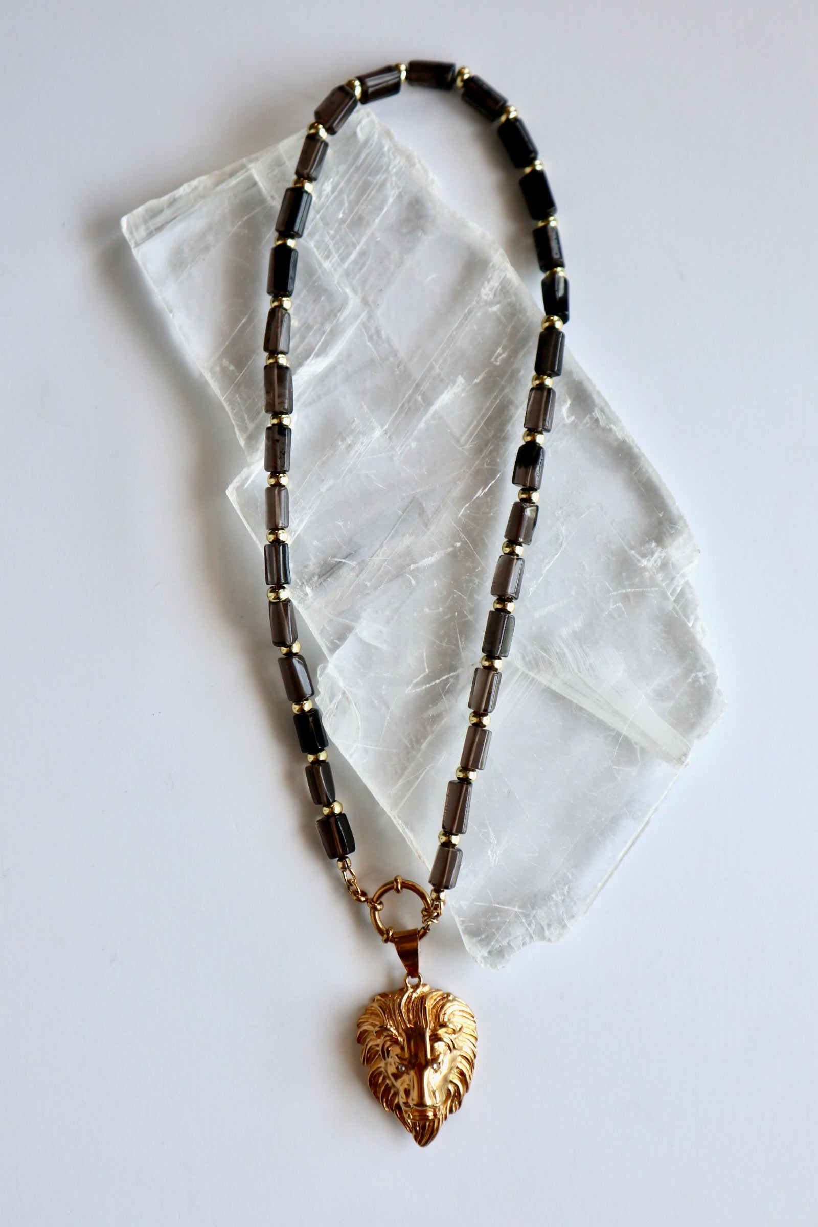 Auréad Designs Leoven necklace laid flat on white selenite slab, showing full strand of faceted smoky quartz cylinder beads with gold spacers, spring ring clasp, and gold lion head pendant with CZ eyes