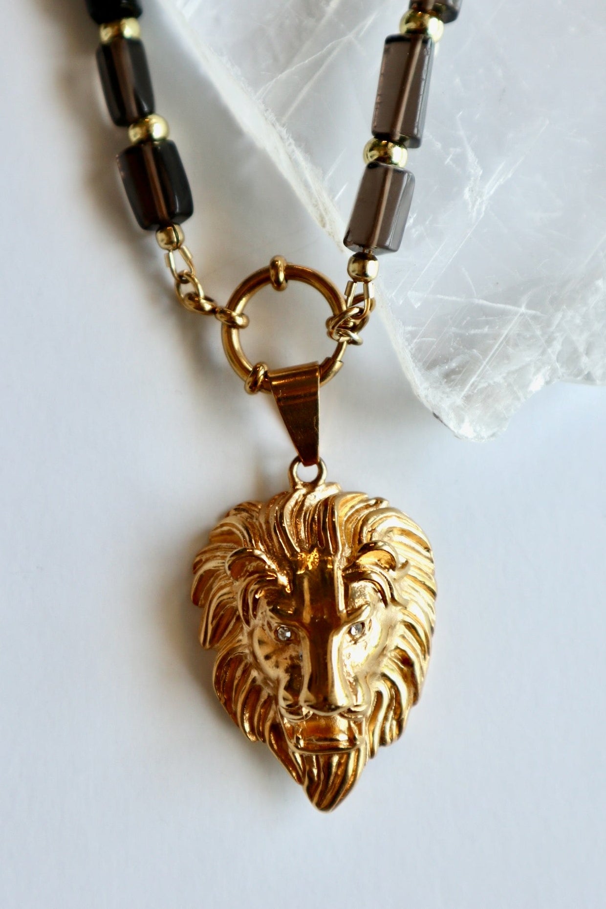 Auréad Designs Leoven necklace on white selenite slab showing gold lion head pendant with CZ eyes and spring ring clasp connecting to smoky quartz cylinder bead strand