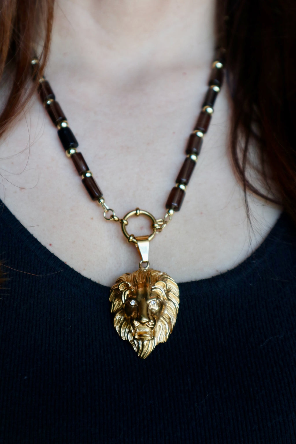 Auréad Designs Leoven necklace worn at collarbone length on a black ribbed top, showing smoky quartz cylinder beads with gold spacers and gold lion head pendant with CZ eyes suspended from spring ring clasp at front

