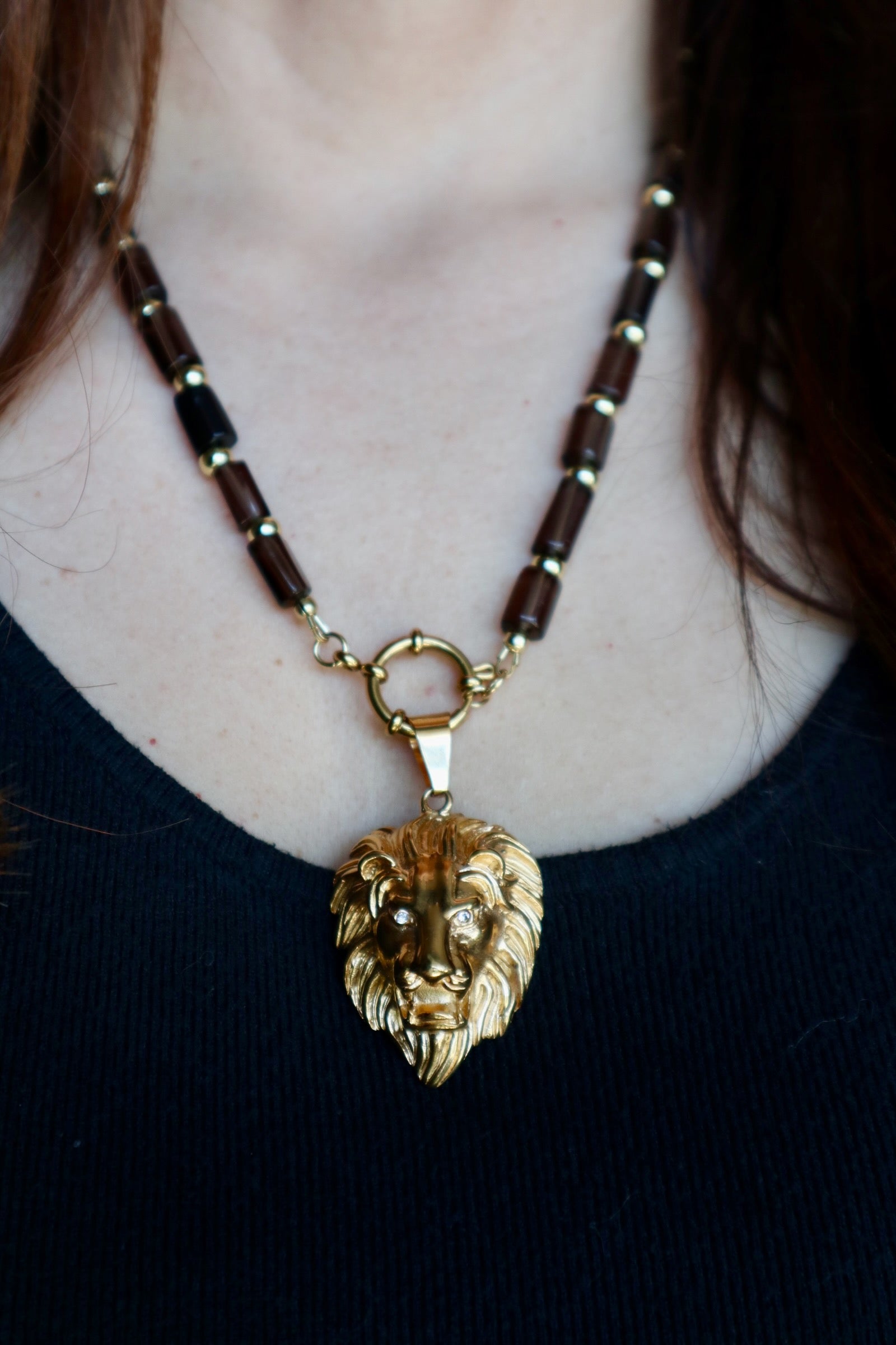 Auréad Designs Leoven necklace worn at collarbone length on a black ribbed top, showing smoky quartz cylinder beads with gold spacers and gold lion head pendant with CZ eyes suspended from spring ring clasp at front

