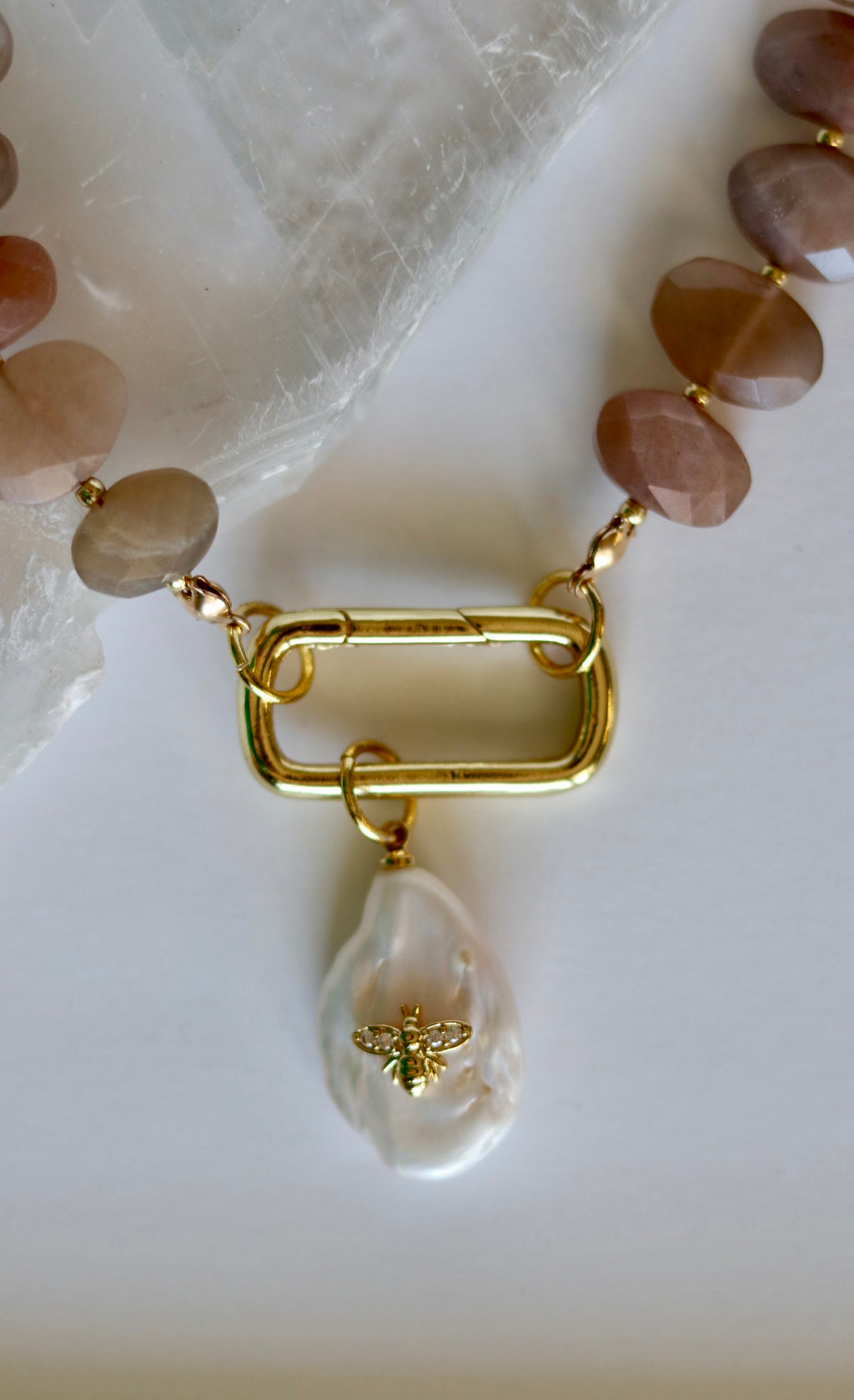 Close-up of the Lucent necklace showing the gold-filled carabiner clasp and removable freshwater pearl charm alongside peach moonstone and gold-filled spacer beads