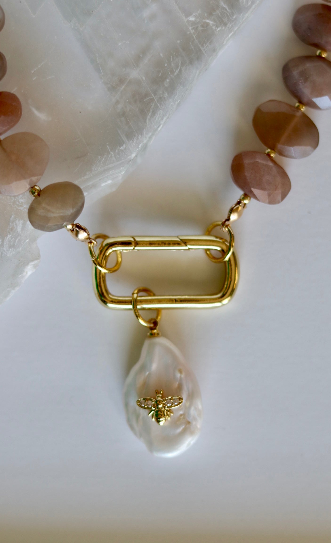 Close-up of the Lucent necklace showing the gold-filled carabiner clasp and removable freshwater pearl charm alongside peach moonstone and gold-filled spacer beads