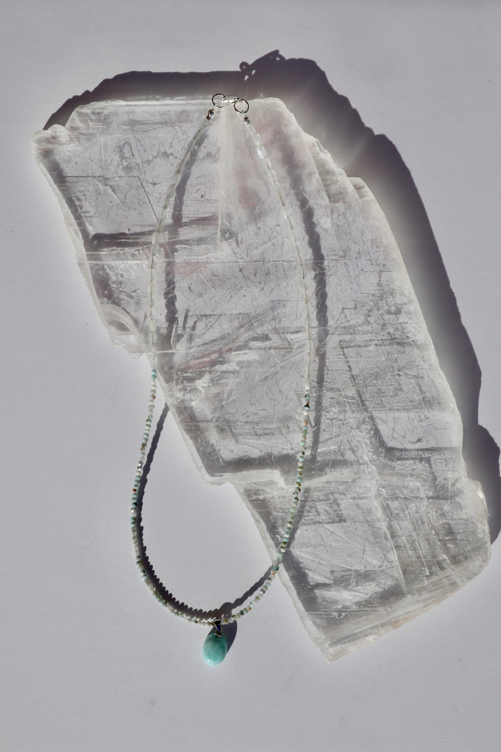 Lunamar necklace by Auréad Designs, featuring micro-faceted larimar beads, flat oval rainbow moonstone beads, and a larimar pendant with a sterling silver lobster clasp, laid flat on a white selenite slab