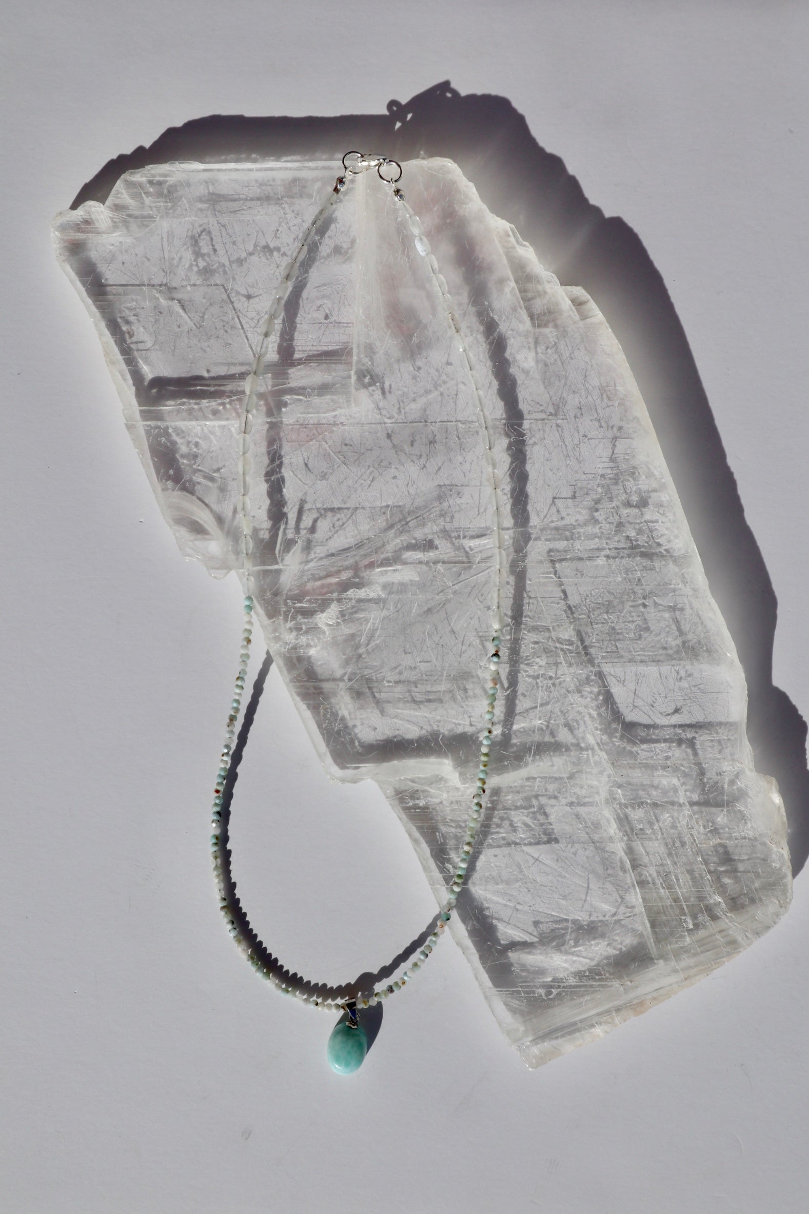 Lunamar necklace by Auréad Designs, featuring micro-faceted larimar beads, flat oval rainbow moonstone beads, and a larimar pendant with a sterling silver lobster clasp, laid flat on a white selenite slab
