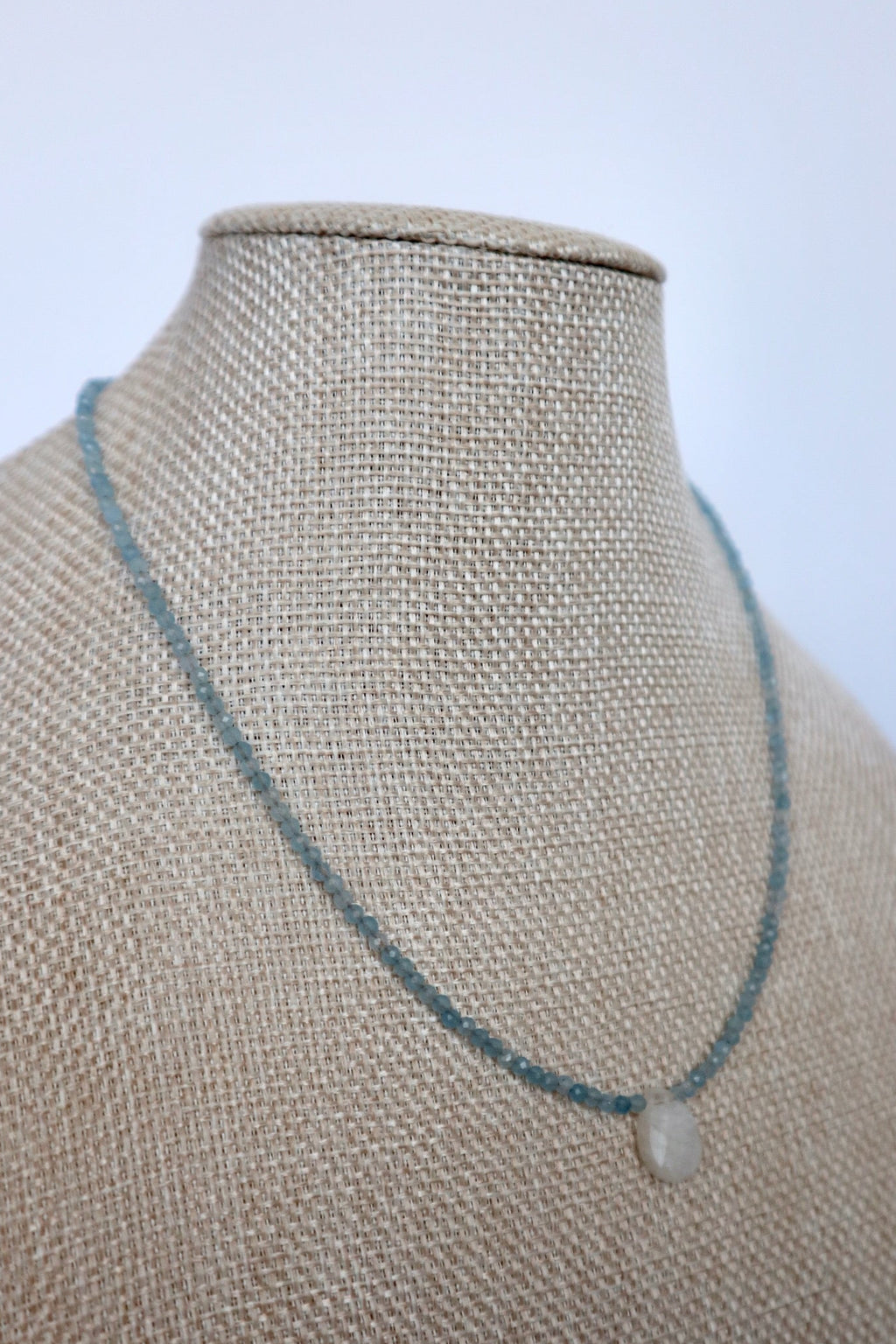 Auréad Designs Marevane aquamarine beaded necklace with rainbow moonstone teardrop pendant displayed at an angle on a linen bust form, showing drape and 16-inch length.