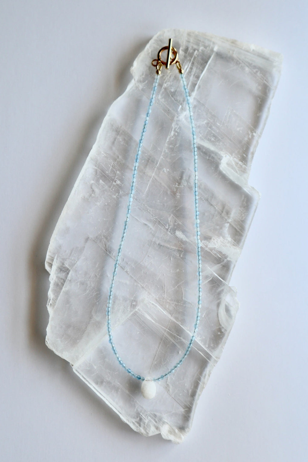 Auréad Designs Marevane necklace with faceted aquamarine beads and rainbow moonstone teardrop pendant with gold toggle clasp, flat lay on white selenite slab.