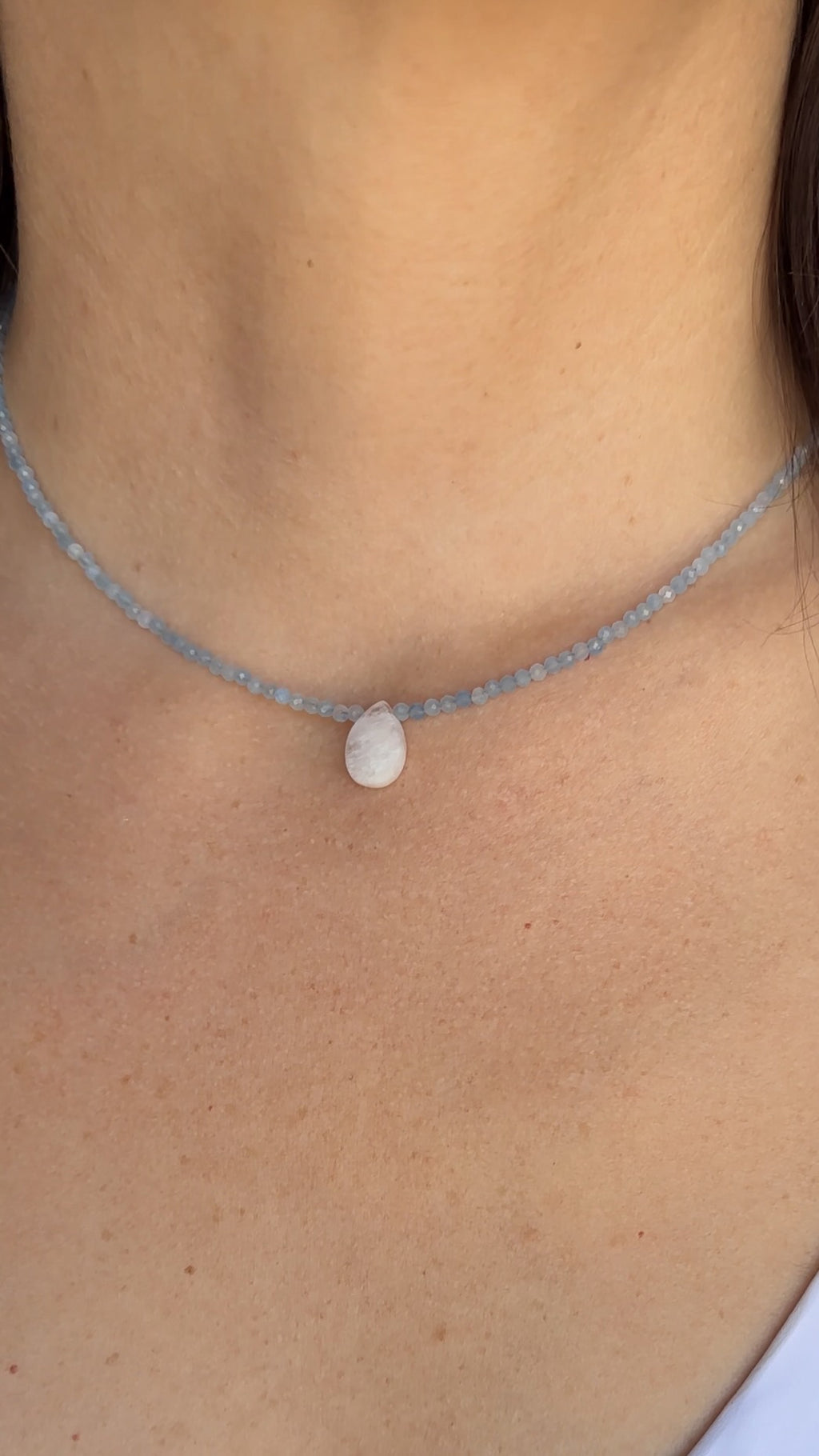Auréad Designs Marevane aquamarine beaded necklace with rainbow moonstone teardrop pendant worn at the collarbone over a white top.
