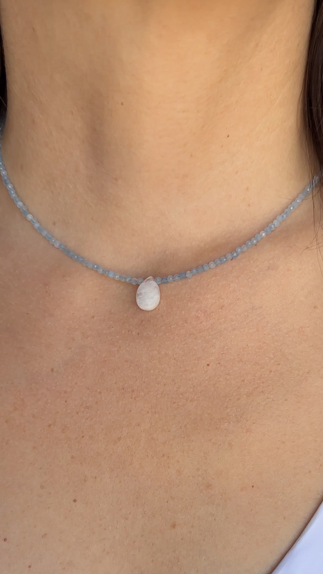 Auréad Designs Marevane aquamarine beaded necklace with rainbow moonstone teardrop pendant worn at the collarbone over a white top.