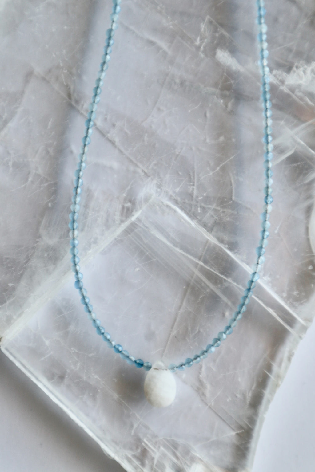 Auréad Designs Marevane necklace showing faceted aquamarine bead strand and rainbow moonstone teardrop pendant, flat lay on white selenite slab.