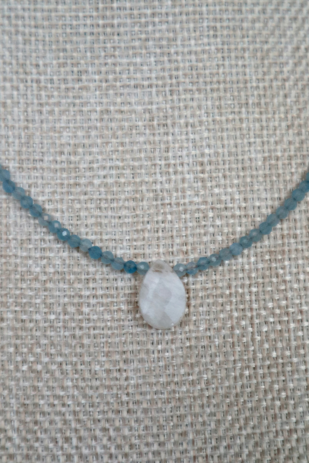 Close-up of Auréad Designs Marevane rainbow moonstone teardrop pendant centered on faceted aquamarine bead strand, on a linen bust form.