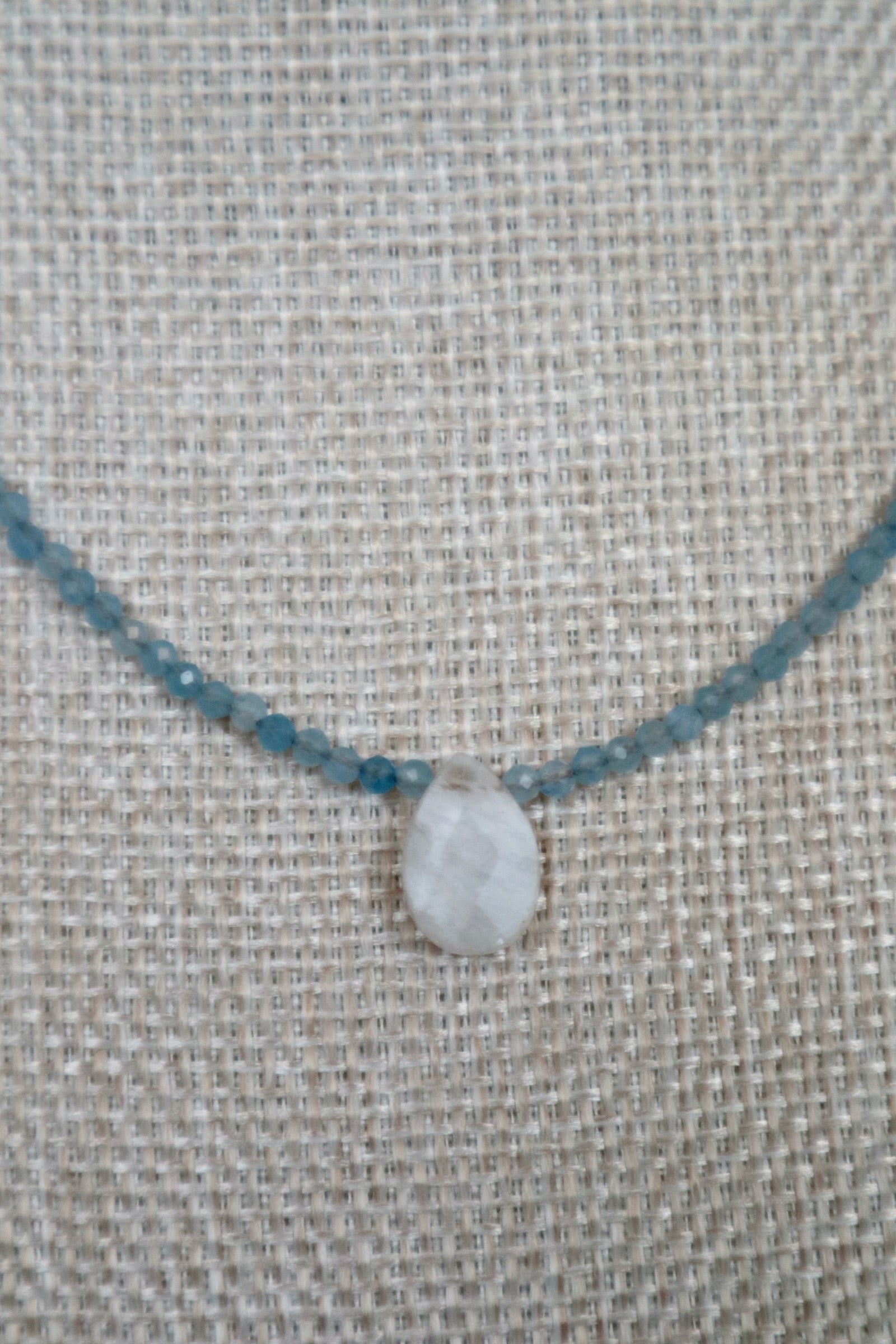Close-up of Auréad Designs Marevane rainbow moonstone teardrop pendant centered on faceted aquamarine bead strand, on a linen bust form.