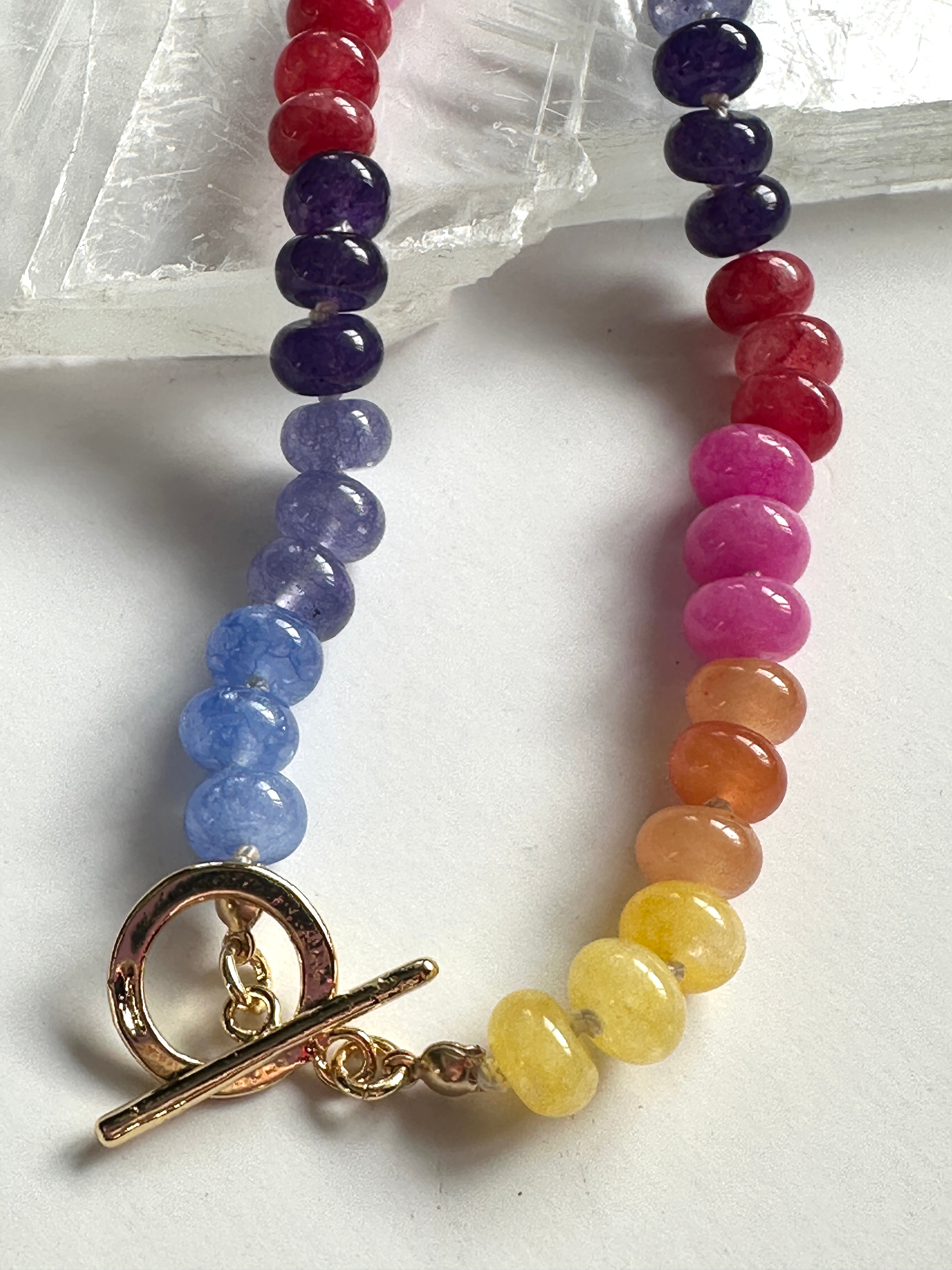 Close-up of the Maribelle necklace on a white selenite slab, showing the gold-filled toggle clasp and the rainbow gradient of candy jade beads transitioning from yellow through orange, pink, red, purple, and blue