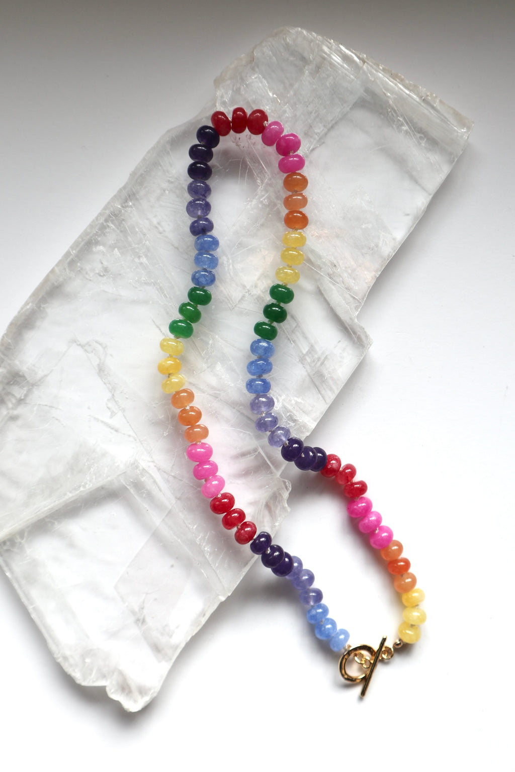 Maribelle necklace by Auréad Designs, featuring colorful candy jade beads on a white silk-knotted strand with a gold-filled toggle clasp, laid flat on a white selenite slab