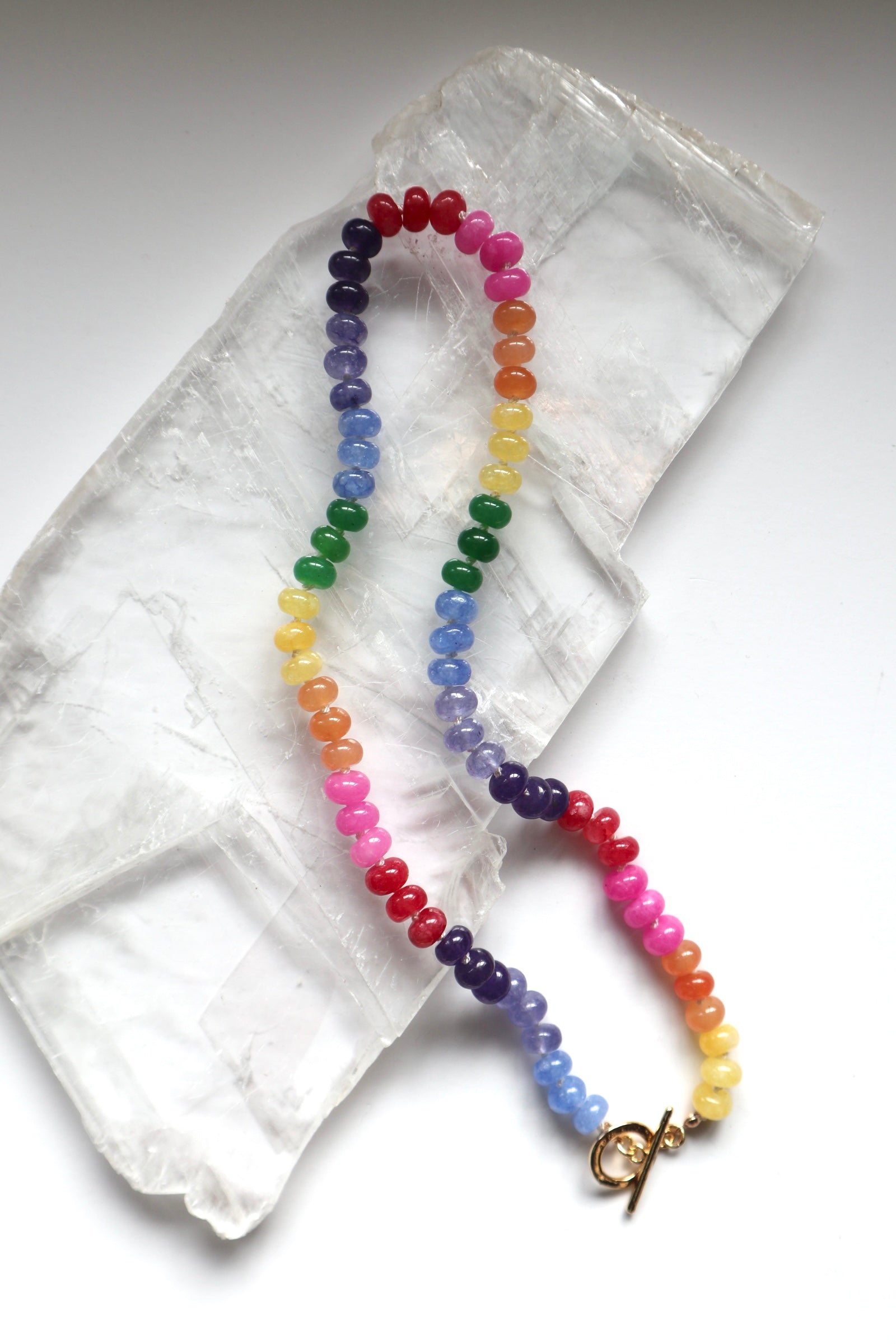 Maribelle necklace by Auréad Designs, featuring colorful candy jade beads on a white silk-knotted strand with a gold-filled toggle clasp, laid flat on a white selenite slab