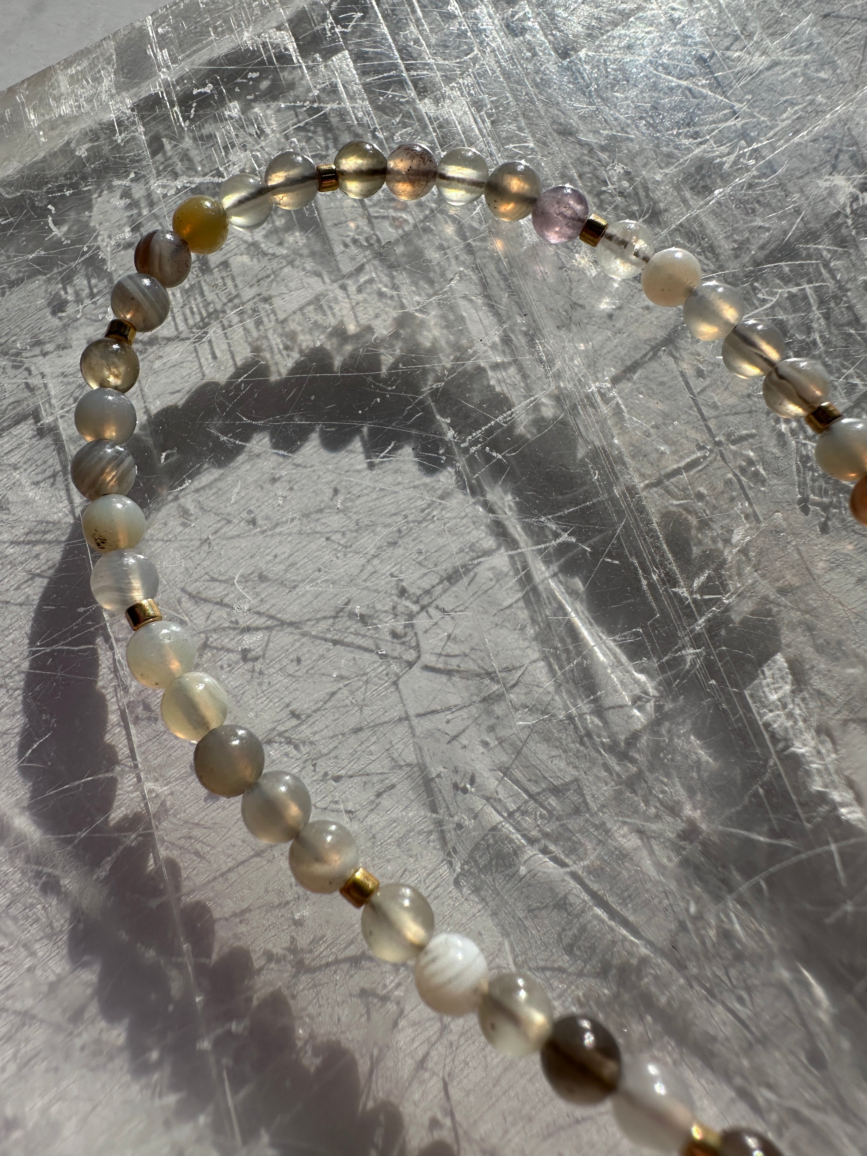 Overhead flat lay of the Marula necklace by Auréad Designs on a white selenite slab, showing the full bead strand and the natural gray, taupe, and warm neutral banding of Botswana agate beads with galvanized gold spacers