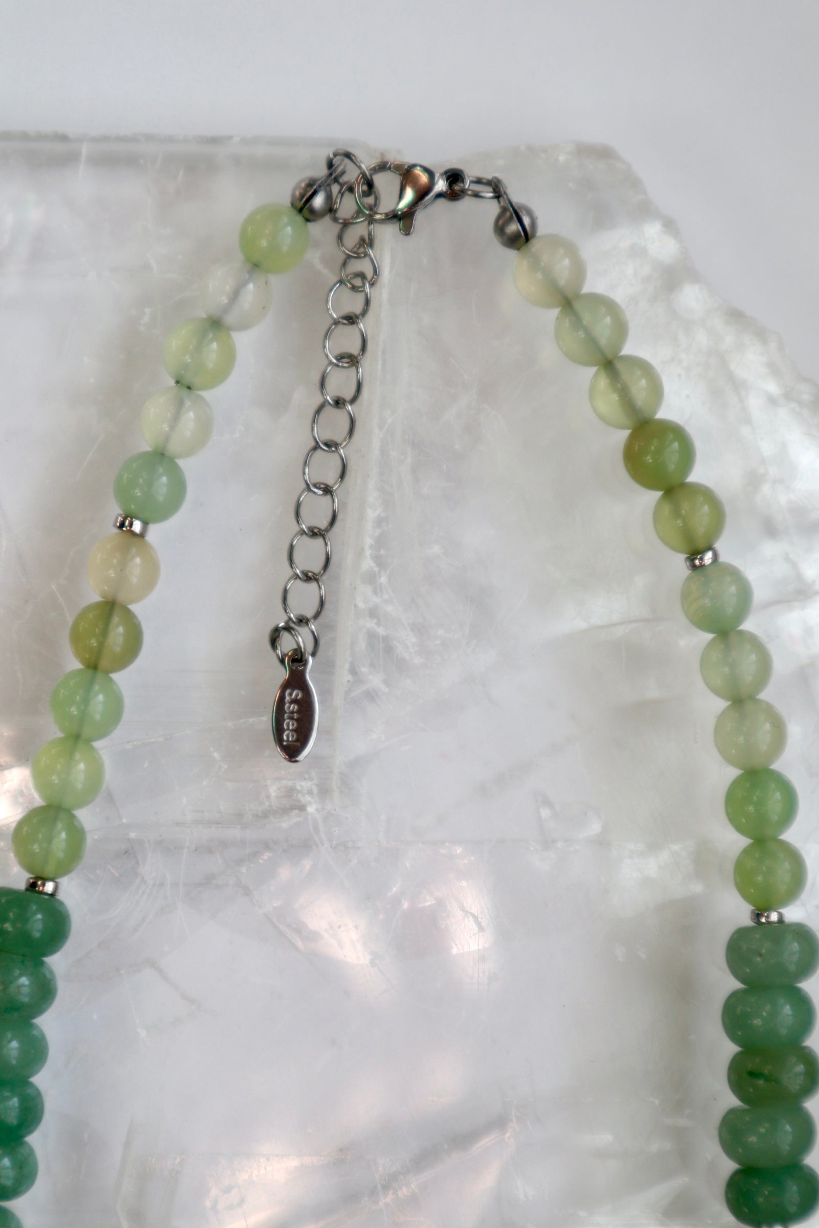 Close-up of the Meadowe necklace on a white selenite slab, showing the stainless steel lobster clasp and extender chain alongside the natural gradient of green jade beads
