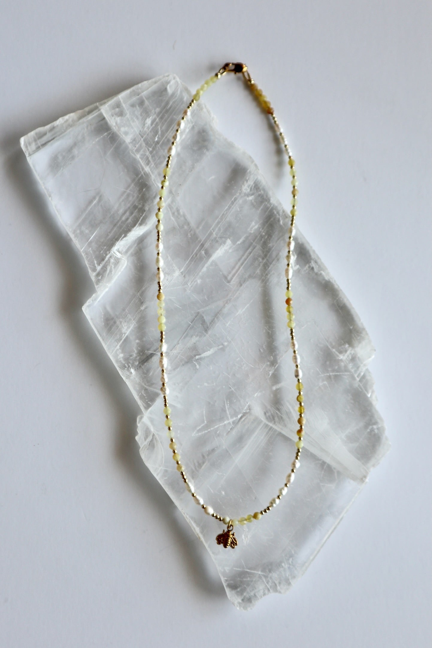 Auréad Designs Melline necklace laid flat on white selenite slab, showing full strand of yellow opal beads, freshwater rice pearls, gold micro spacer beads, and gold bee charm with lobster clasp
