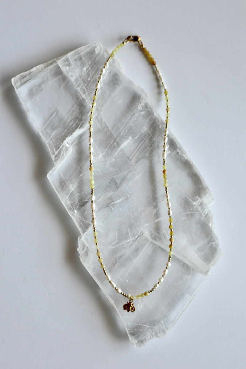 Auréad Designs Melline necklace laid flat on white selenite slab, showing full strand of yellow opal beads, freshwater rice pearls, gold micro spacer beads, and gold bee charm with lobster clasp