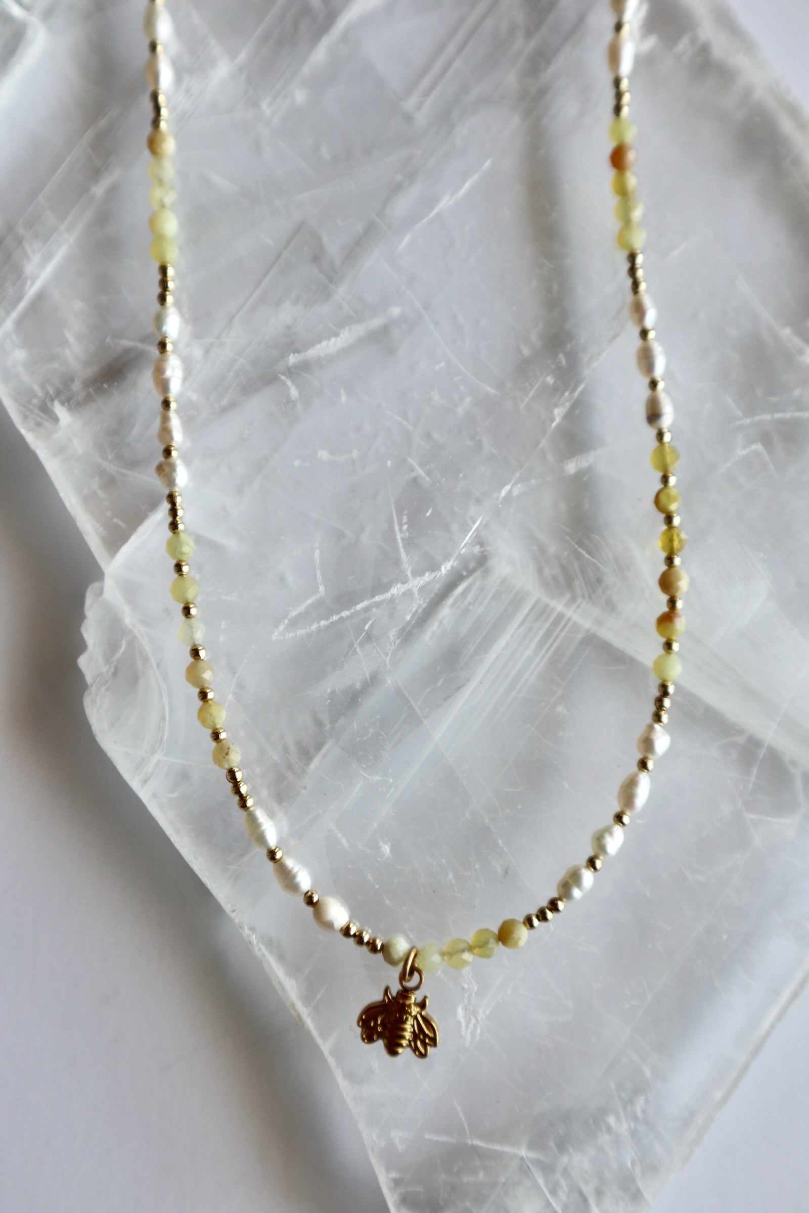 Auréad Designs Melline necklace on white selenite slab showing yellow opal bead coloring, freshwater rice pearls, gold spacers, and dangling gold bee charm at center front