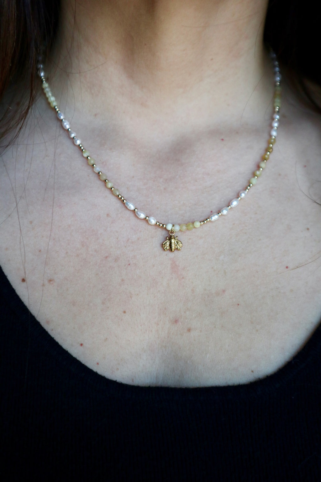 Auréad Designs Melline necklace worn at collarbone length on a black top, showing yellow opal beads, freshwater rice pearls, and gold micro spacers with gold bee charm centered at front against bare skin