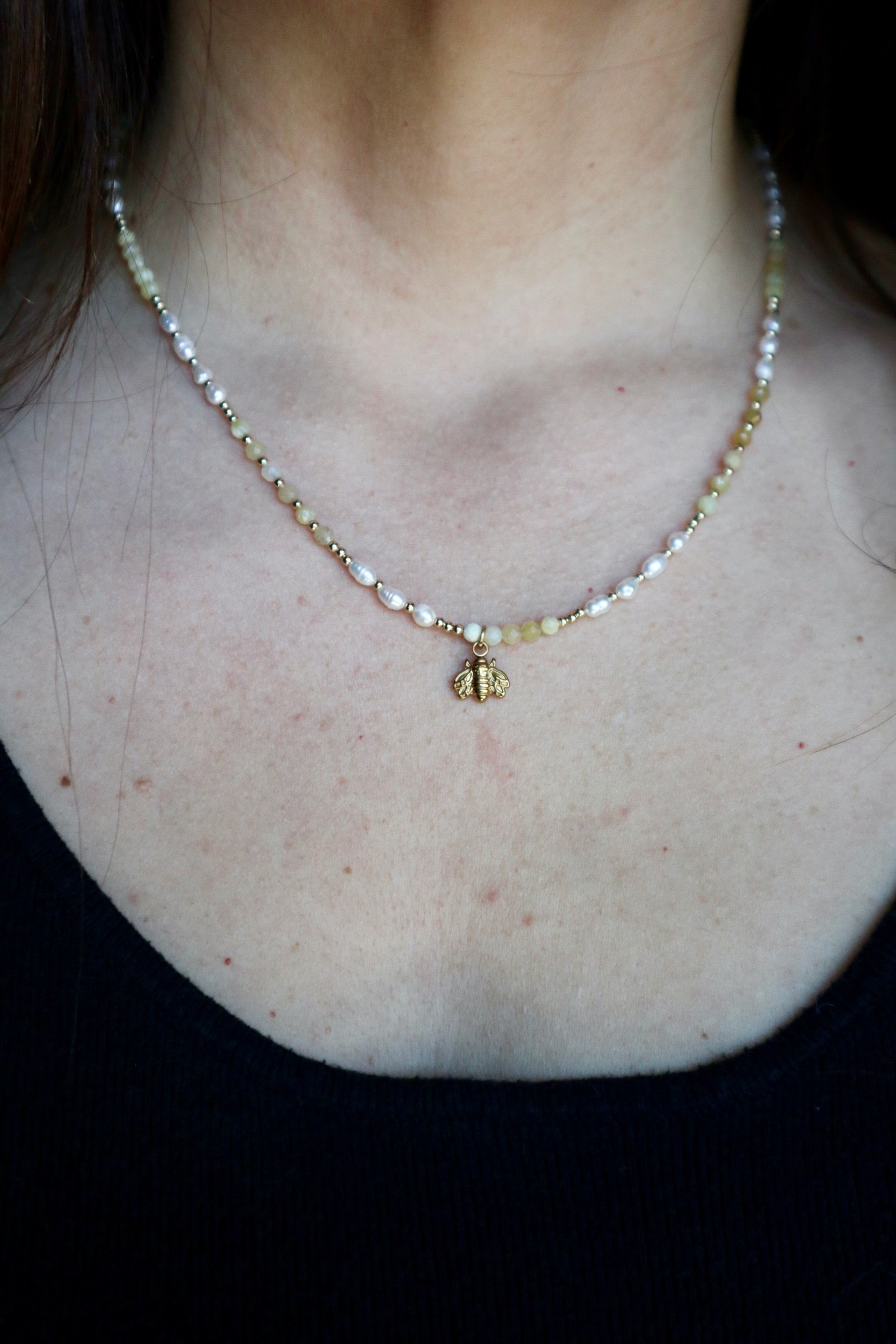 Auréad Designs Melline necklace worn at collarbone length on a black top, showing yellow opal beads, freshwater rice pearls, and gold micro spacers with gold bee charm centered at front against bare skin