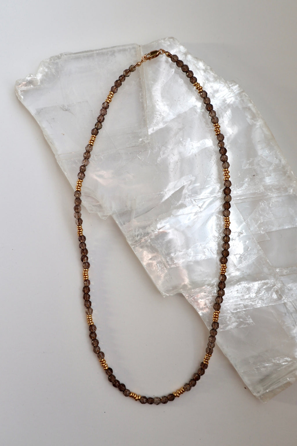 Mira necklace by Auréad Designs, featuring smoky quartz gemstone beads and galvanized gold spacer beads with a 14k gold-filled clasp, laid flat on a white selenite slab