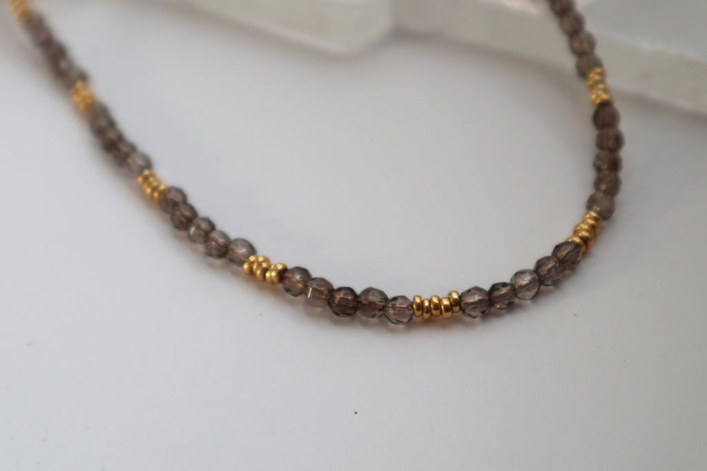 Close-up of the Mira necklace resting on a white selenite slab, showing the translucent smoky quartz beads and galvanized gold spacer beads in detail
