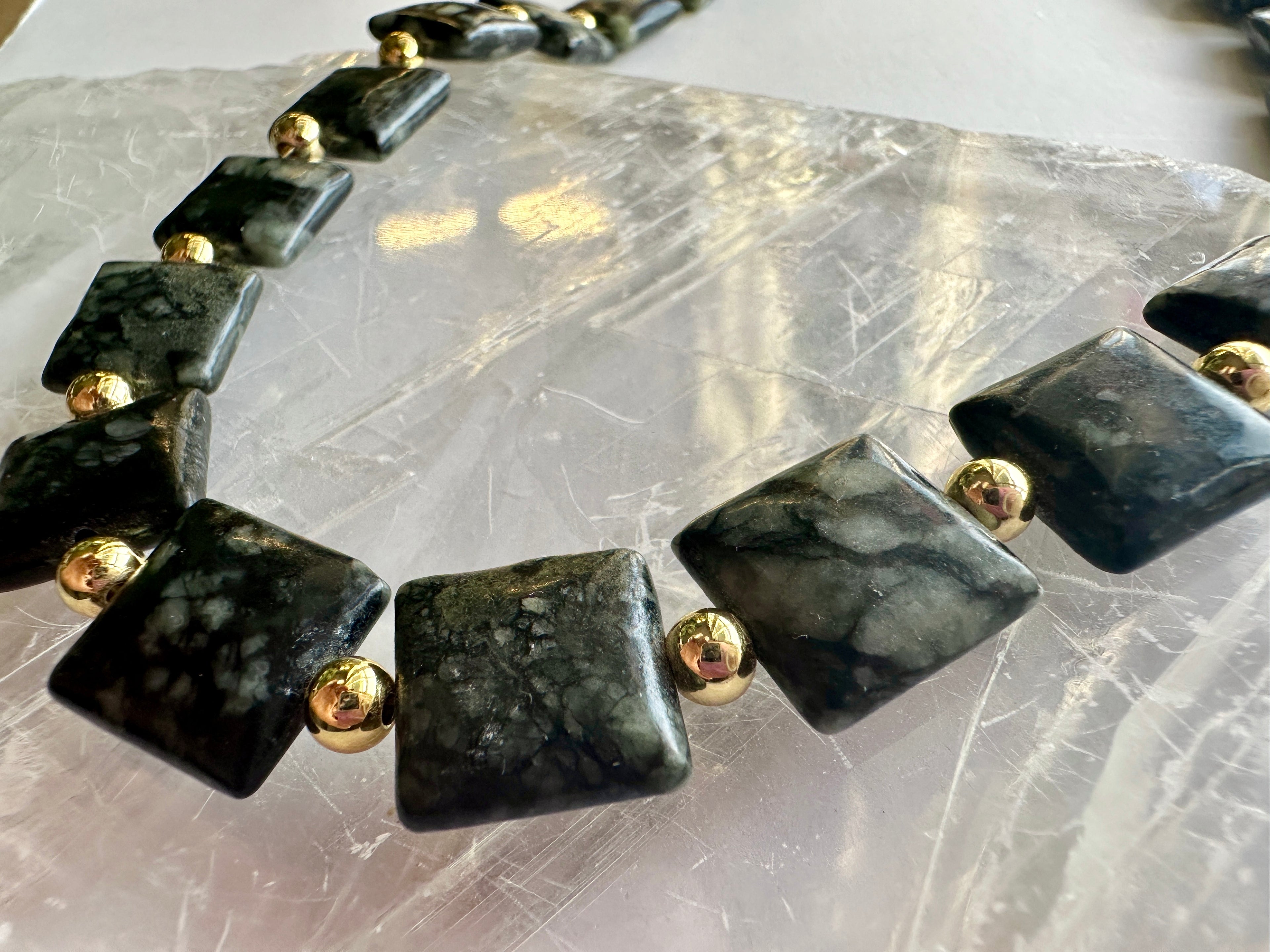 Macro flat lay of the Morenne necklace by Auréad Designs on a white selenite slab, showing the full arrangement of Picasso serpentine pillow beads and gold-filled round spacers with their natural olive, charcoal, and grey patterning