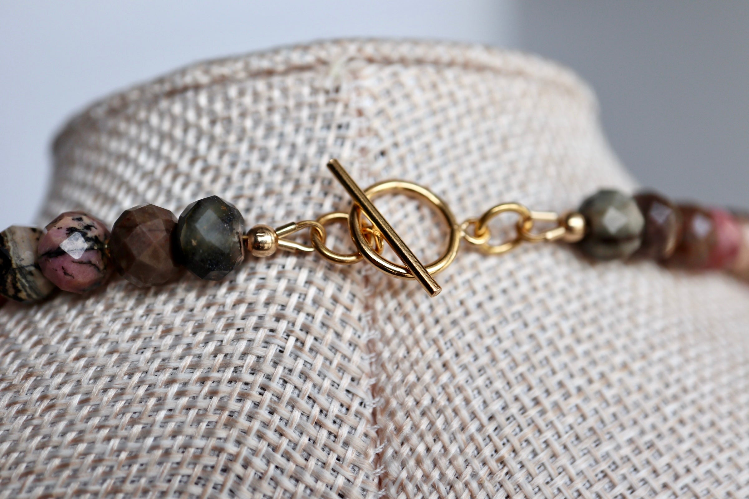 Close-up of the Noirrose necklace on a linen bust form, highlighting the 14k gold-filled toggle clasp and the natural deep rose and black veining of the faceted rhodonite rondelle beads