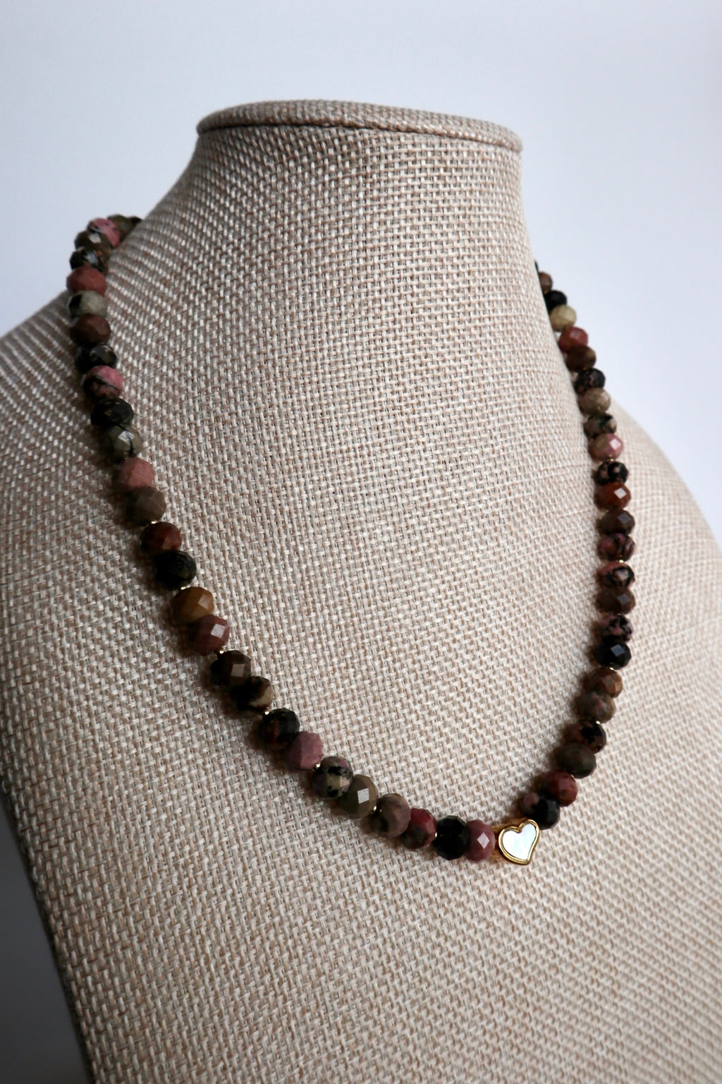 Noirrose necklace by Auréad Designs displayed on a linen bust form, showing the full strand of faceted rhodonite rondelle beads with the gold iridescent heart charm centered at the front