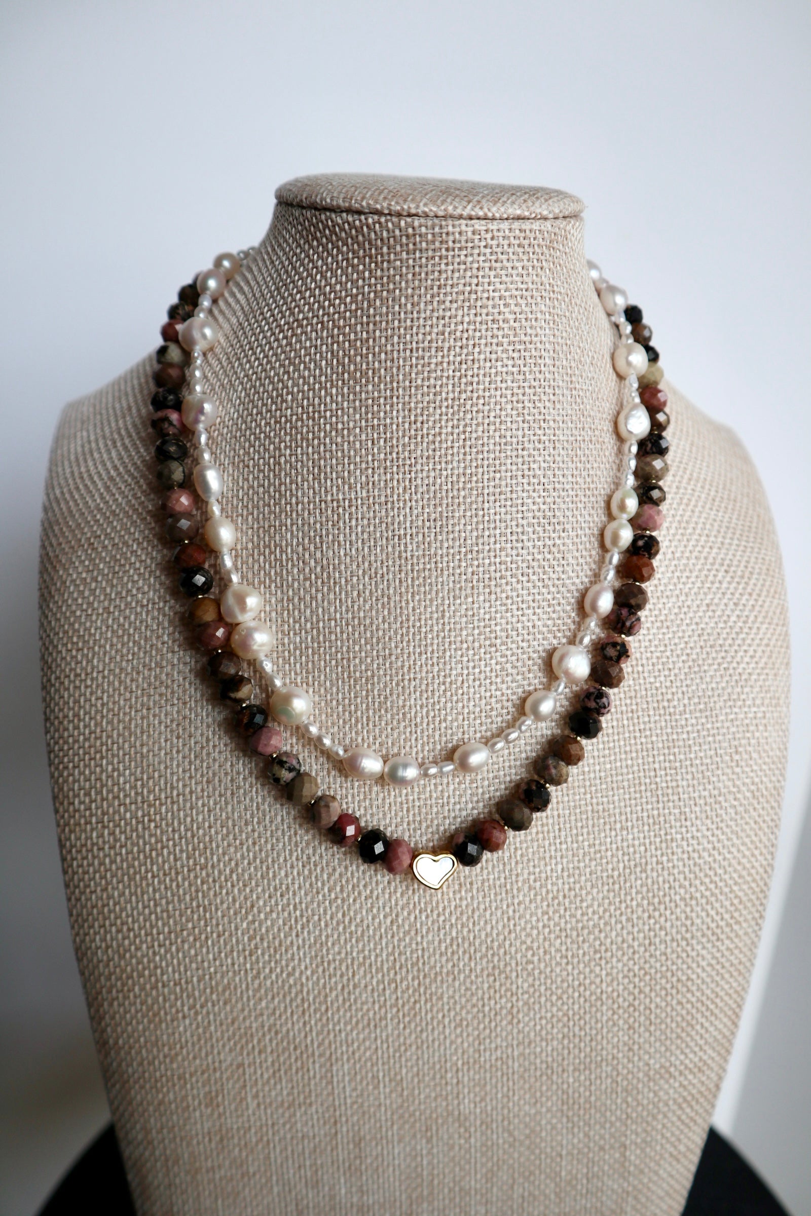 Noirrose necklace by Auréad Designs displayed on a linen bust form, showing the full strand of faceted rhodonite rondelle beads and gold heart charm with iridescent inlay