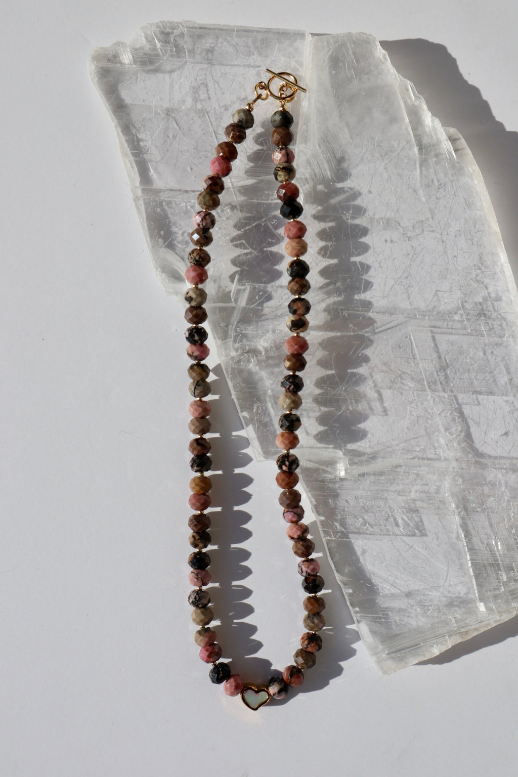 Noirrose necklace by Auréad Designs, featuring faceted rhodonite rondelle beads, a gold stainless steel heart charm with iridescent inlay, and a 14k gold-filled toggle clasp, laid flat on a white selenite slab