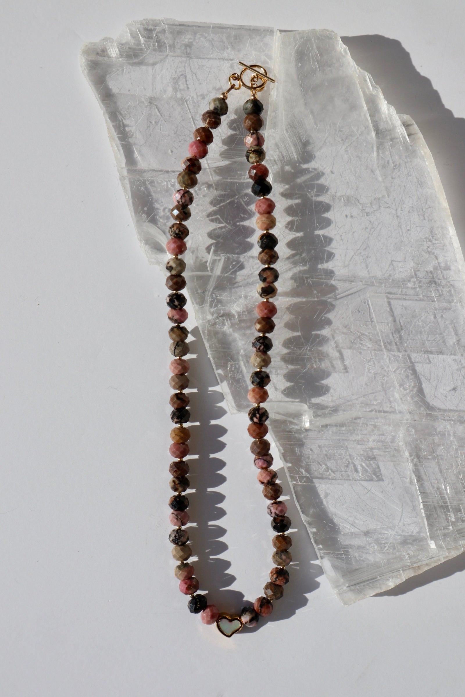 Noirrose necklace by Auréad Designs, featuring faceted rhodonite rondelle beads, a gold stainless steel heart charm with iridescent inlay, and a 14k gold-filled toggle clasp, laid flat on a white selenite slab