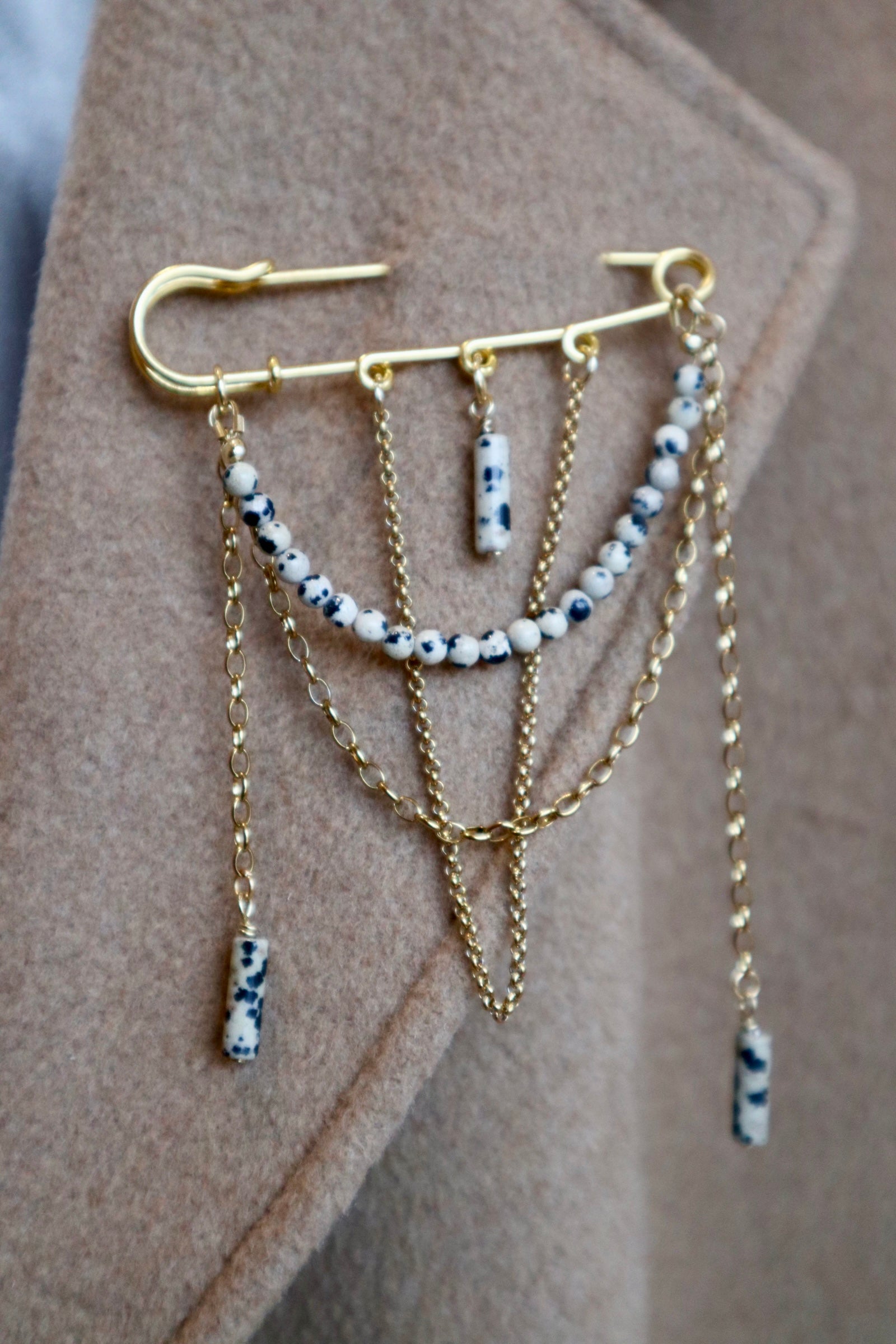 Perrin pin brooch by Auréad Designs worn on a camel coat, showing the full design with layered gold stainless steel chains, Dalmatian jasper round beads, and dangling jasper cylinder drops