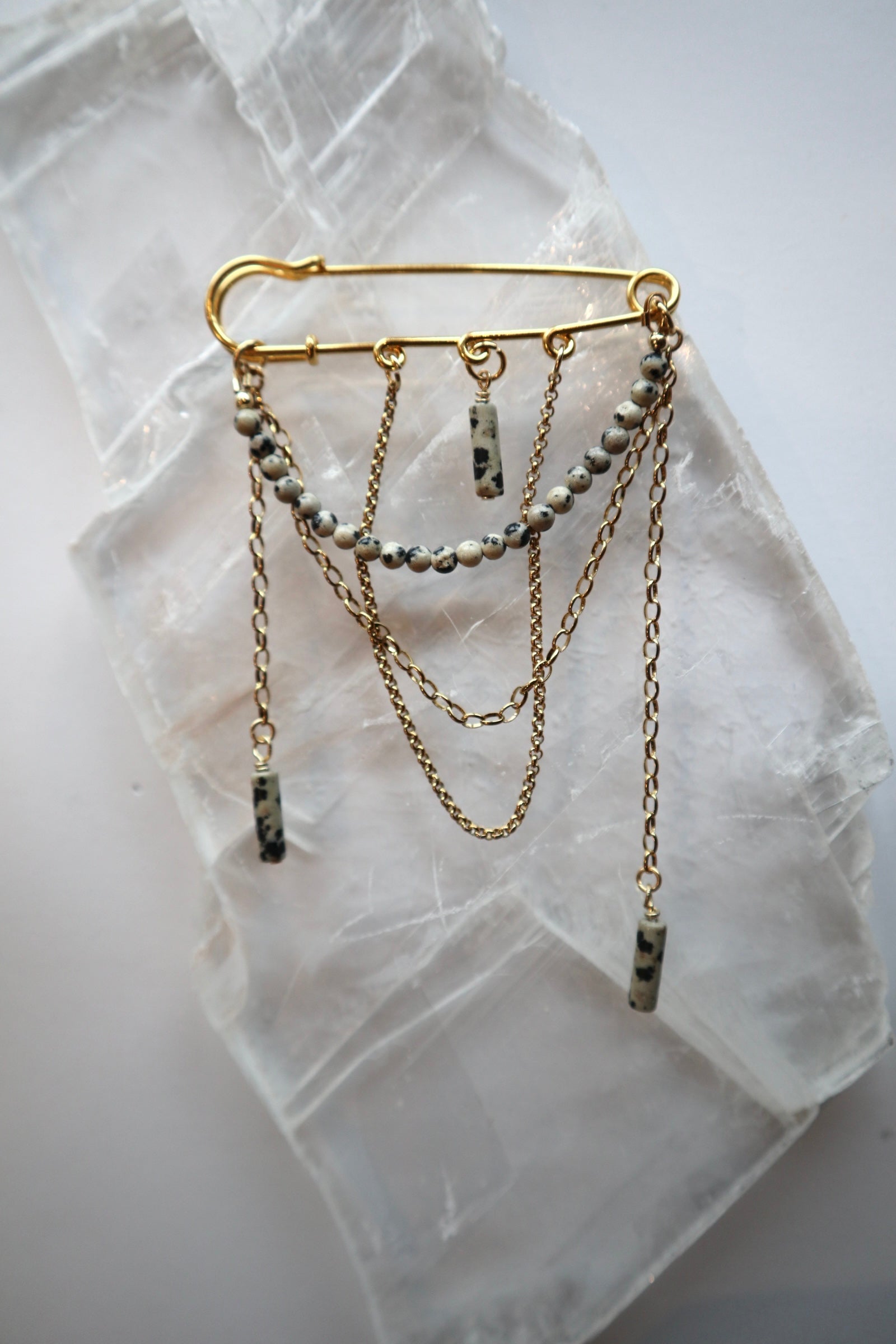 Perrin pin brooch by Auréad Designs, featuring Dalmatian jasper beads and layered gold stainless steel rolo and cable chains, laid flat on a white selenite slab