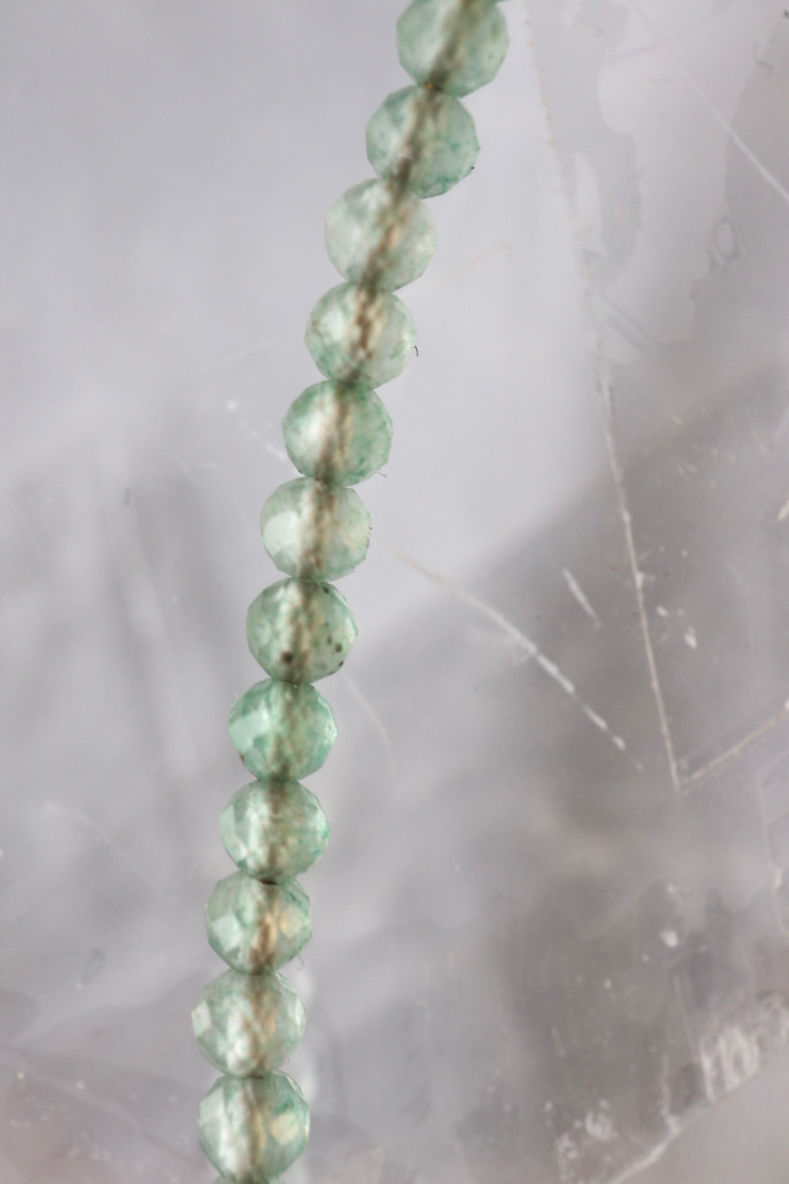 Macro detail of 2mm faceted green aventurine beads from the Auréad Designs Primelle necklace against a selenite slab background, showing natural variation in color and translucency