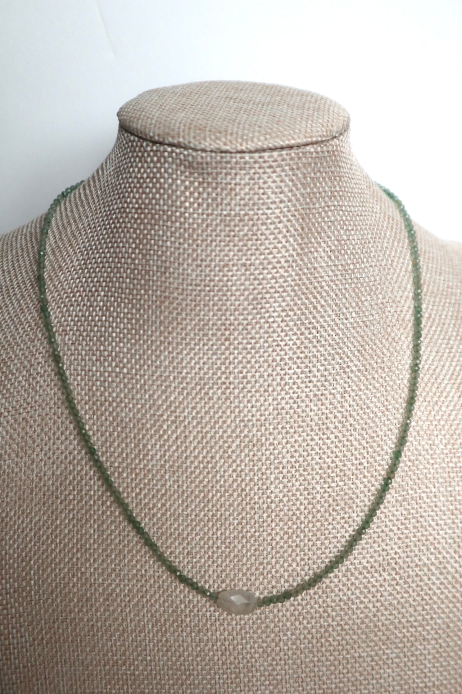 Auréad Designs Primelle necklace displayed on a linen bust form showing full strand of 2mm faceted green aventurine beads with moonstone focal bead at center