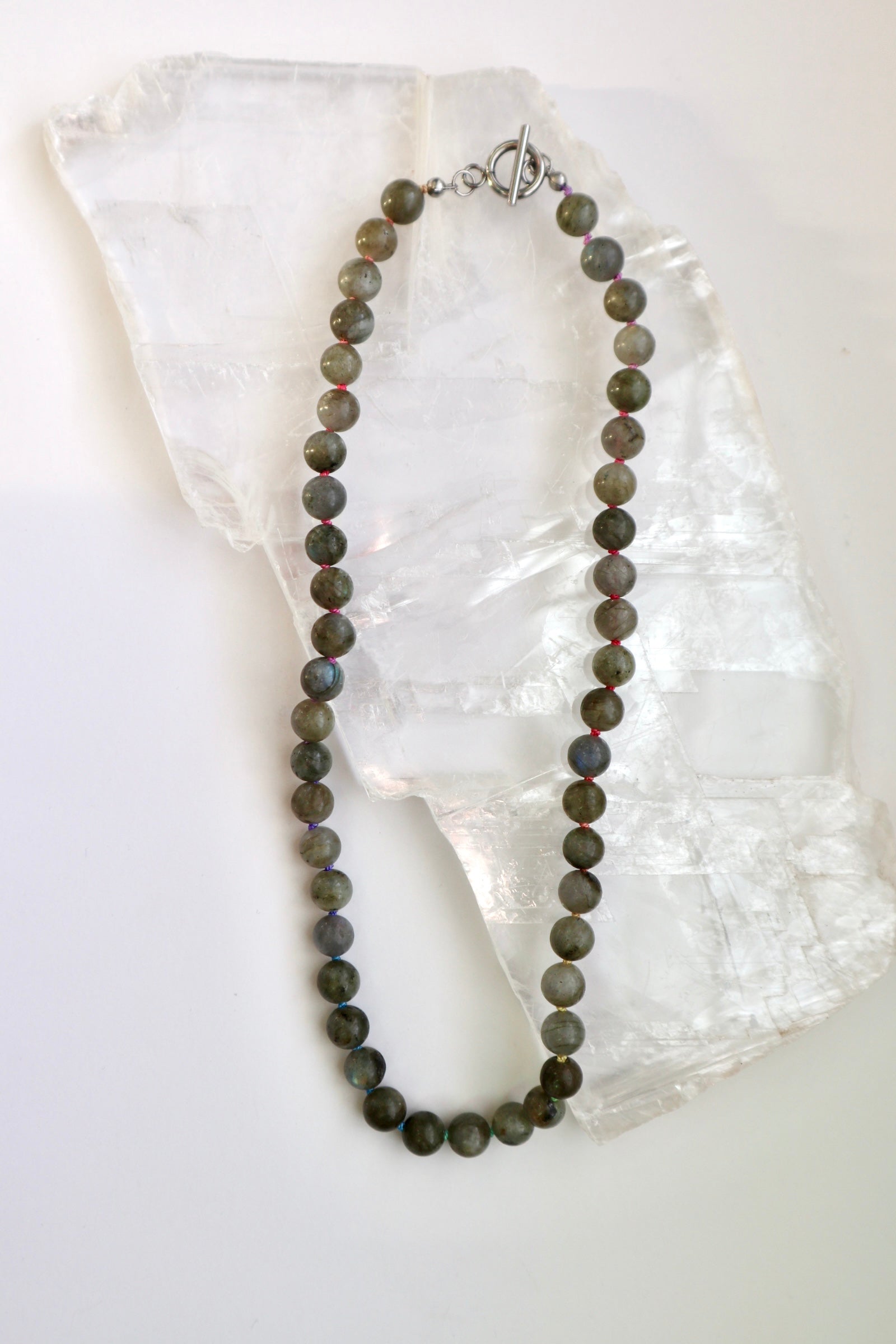 Prism necklace by Auréad Designs, featuring round labradorite gemstone beads on a multicolor ombré silk cord with a stainless steel clasp, laid flat on a white selenite slab