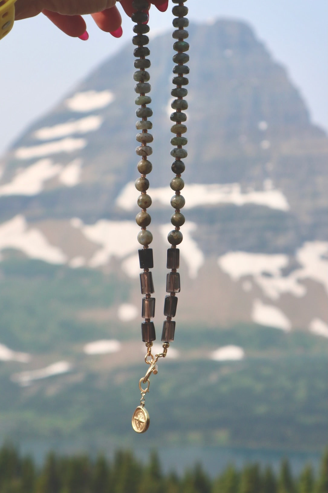 Ridge necklace by Auréad Designs held against a snow-capped mountain peak at Logan Pass, Glacier National Park, with labradorite rondelles, smoky quartz tubes, and a gold compass charm toggle clasp