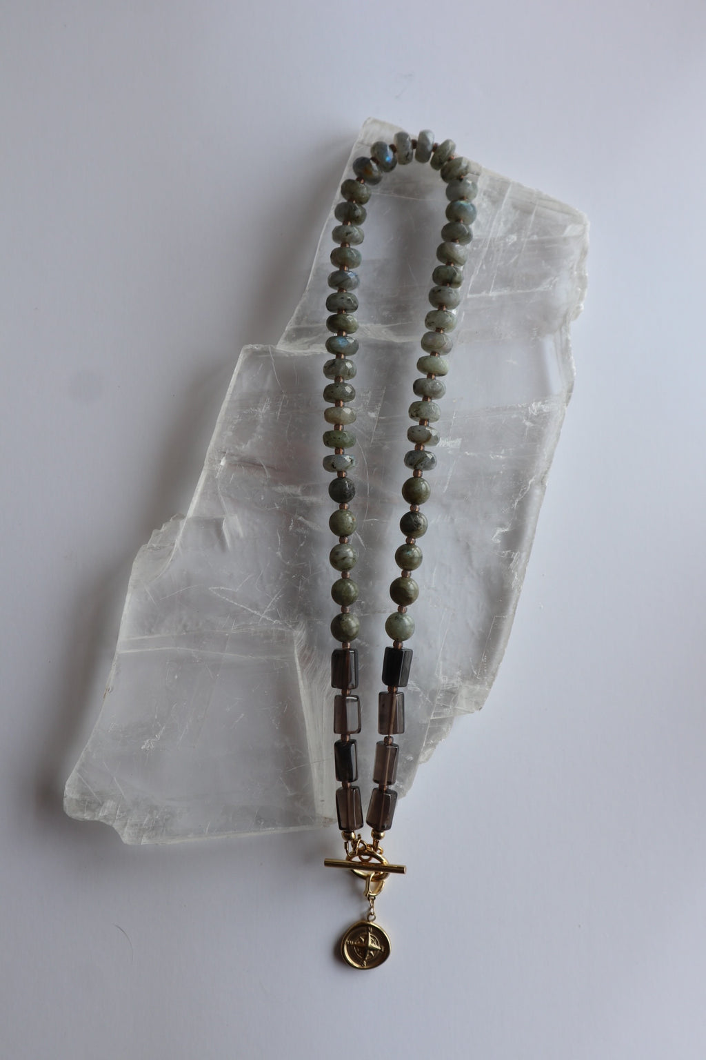 Ridge necklace by Auréad Designs laid flat on a selenite slab, showing the full length with labradorite rondelles, smoky quartz tubes, and gold compass charm toggle clasp