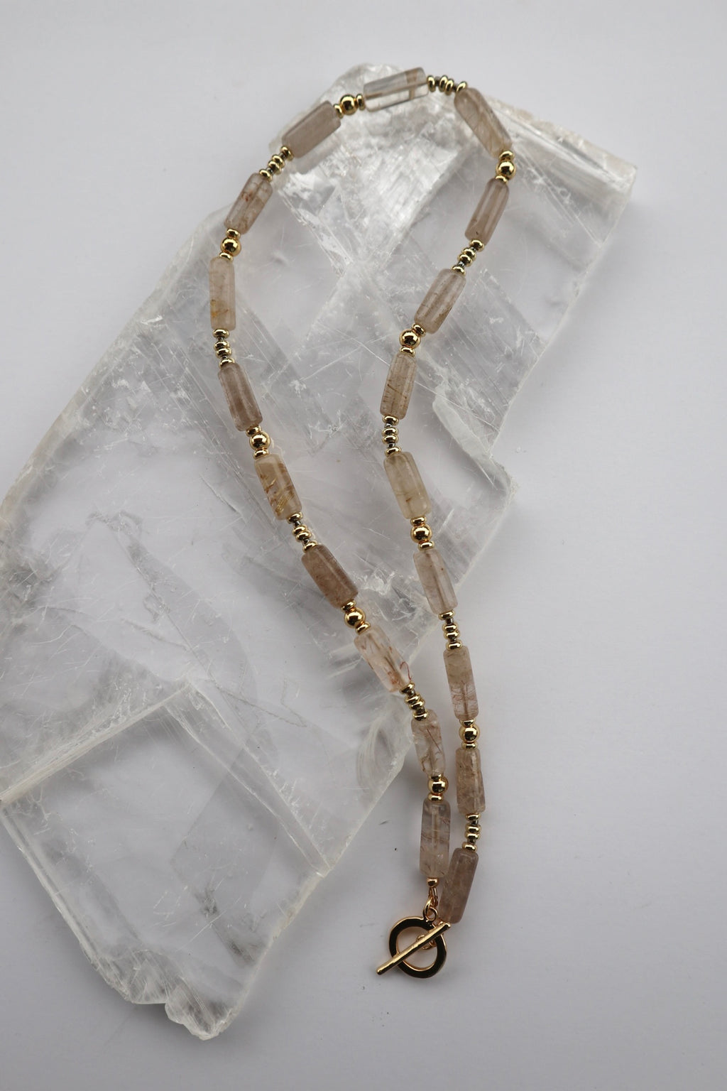 Rutila necklace by Auréad Designs, featuring rutilated quartz cylinder beads, galvanized gold spacer beads, and dark gray glass seed beads with a gold-filled toggle clasp, laid flat on a white selenite slab