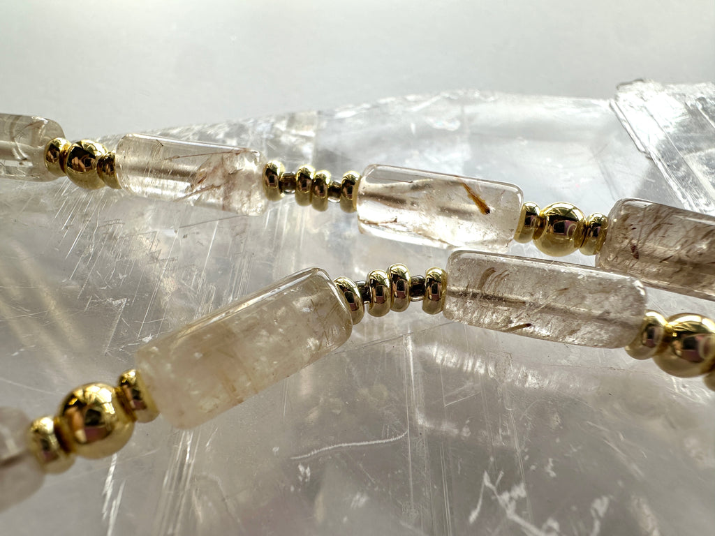 Macro close-up of the Rutila necklace by Auréad Designs resting on a white selenite slab, showing the natural golden rutile inclusions within the clear quartz cylinder beads and galvanized gold spacers