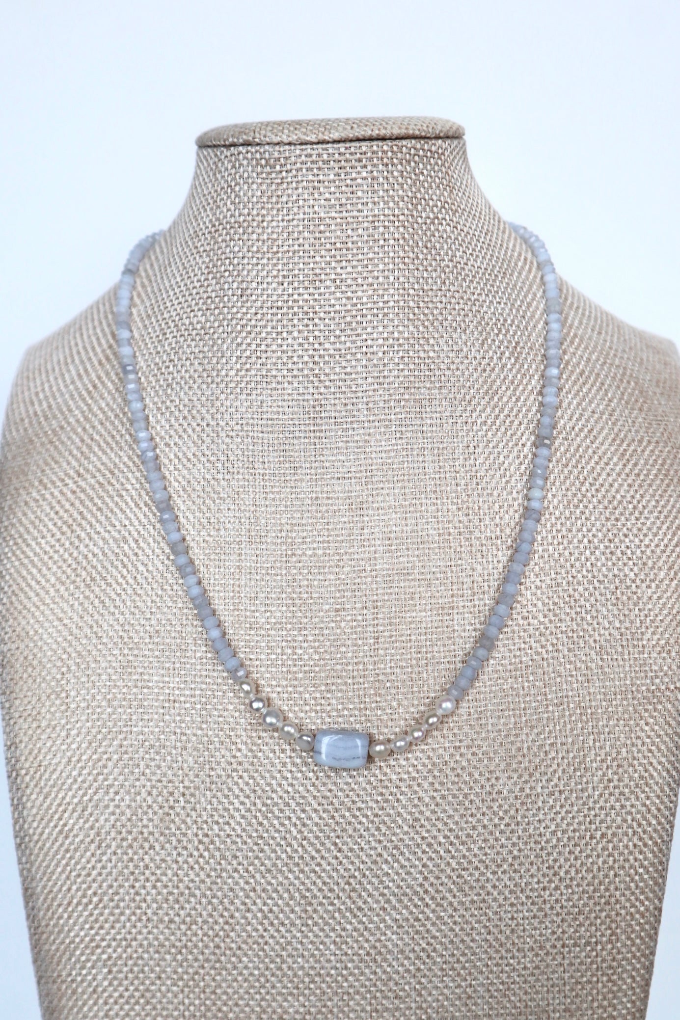 Sky necklace by Auréad Designs on a linen bust, front view, featuring blue lace agate rondelles, freshwater pearl accents, and a rectangular blue lace agate focal bead with a gold toggle clasp