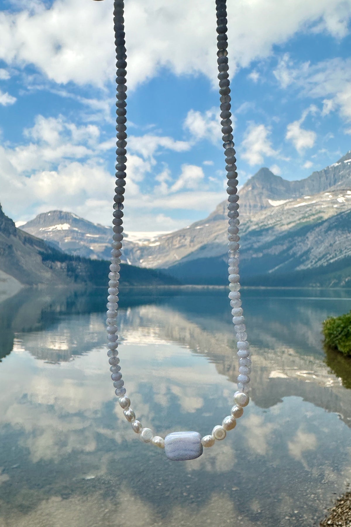 Sky necklace by Auréad Designs held against a glacially-fed mountain lake on the Icefields Parkway, with blue sky and snow-capped peaks reflected in the still water below