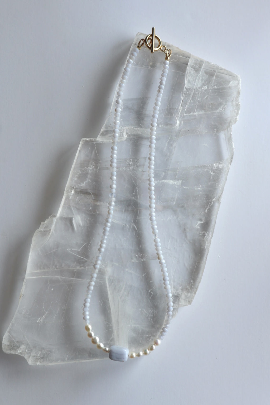 Sky necklace by Auréad Designs laid flat on a selenite slab, showing the full length with blue lace agate rondelles, freshwater pearl accents, rectangular focal bead, and gold toggle clasp